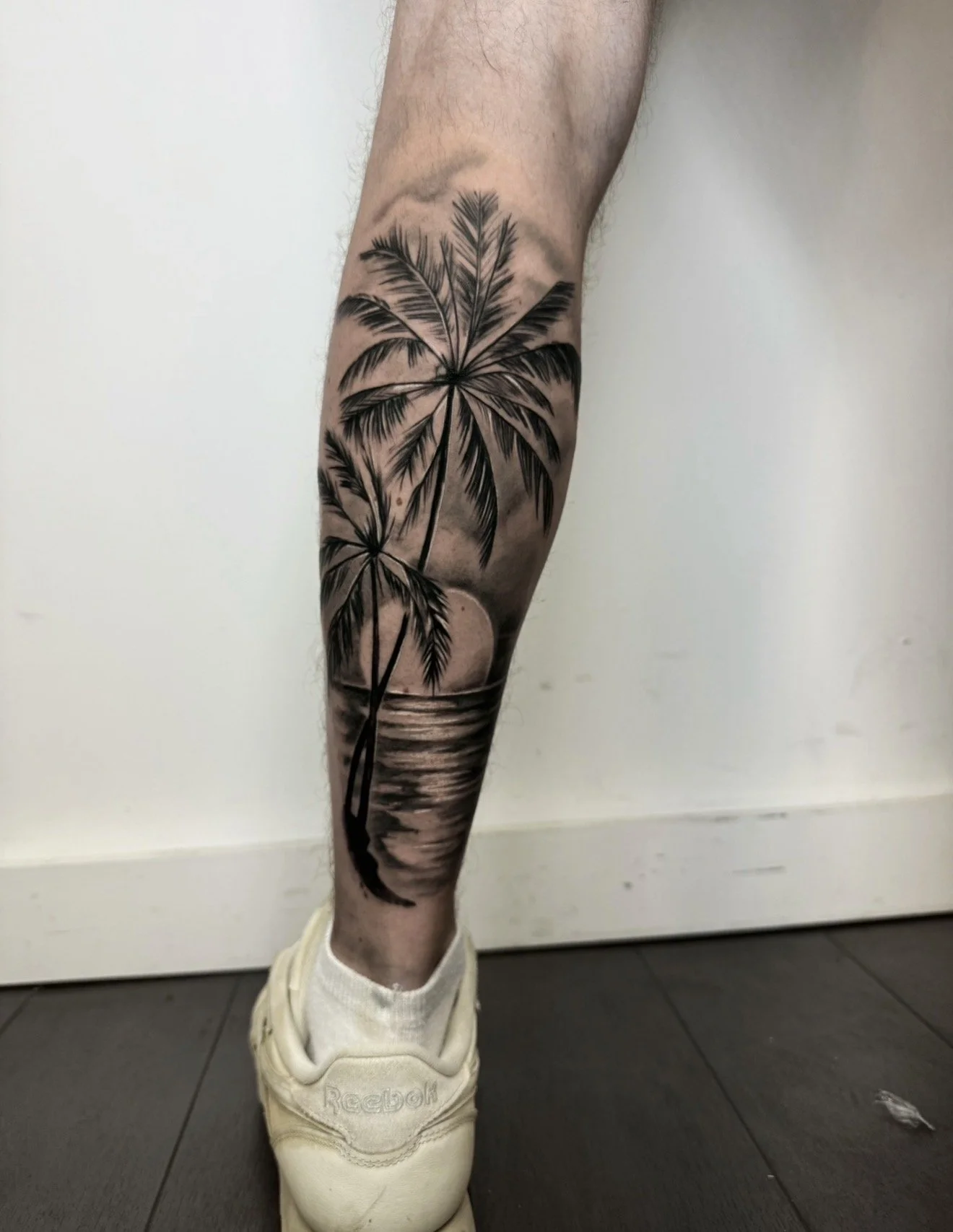 Tattoo of palm trees and a sunset on a person's lower leg, with a white Reebok shoe and white sock visible, standing on a dark tiled floor against a plain white wall.