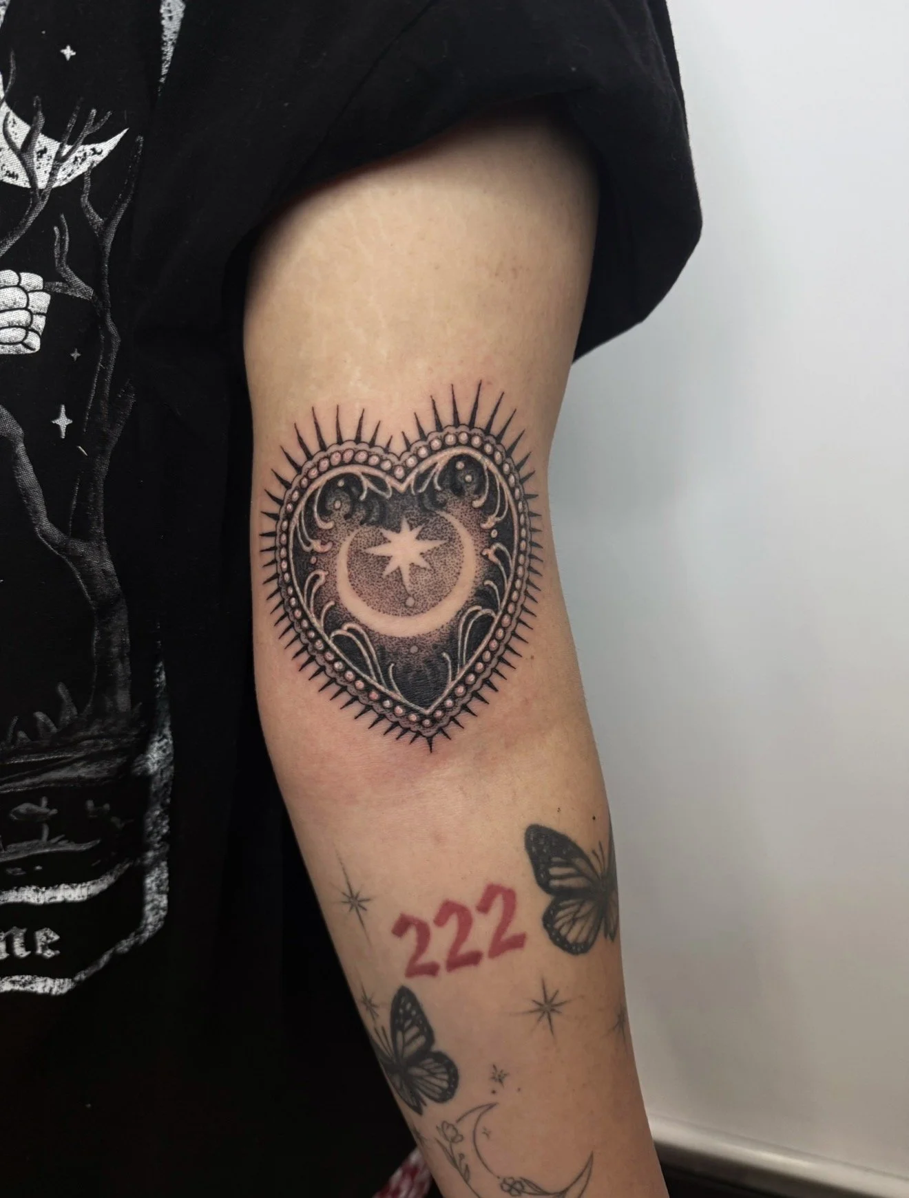 Tattoo of a heart-shaped design with a crescent moon and stars inside, surrounded by decorative lines and dots, on a person's upper arm.