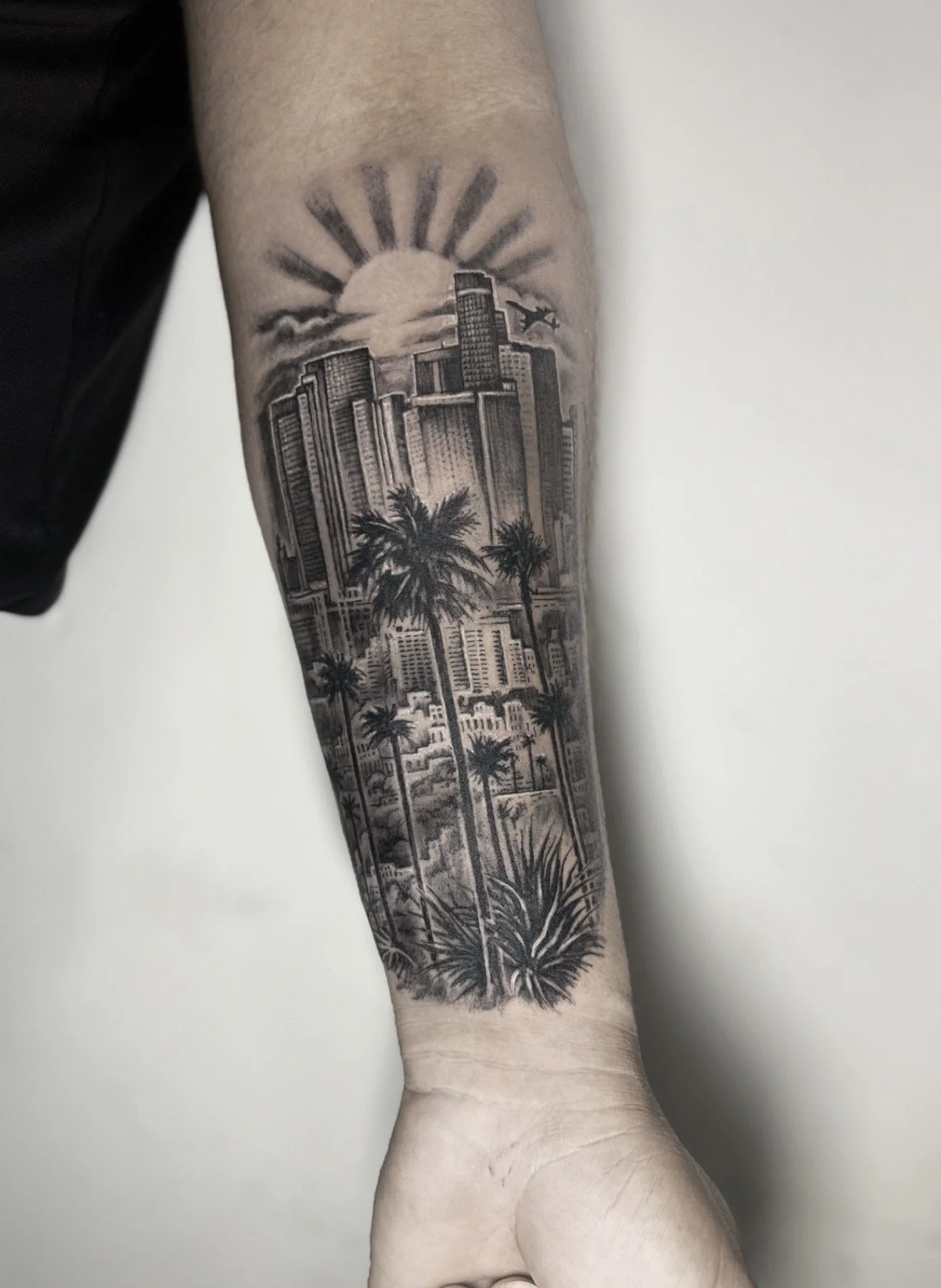 Tattoo of a city skyline with tall buildings, palm trees, and a sunset with rays on a person's forearm.