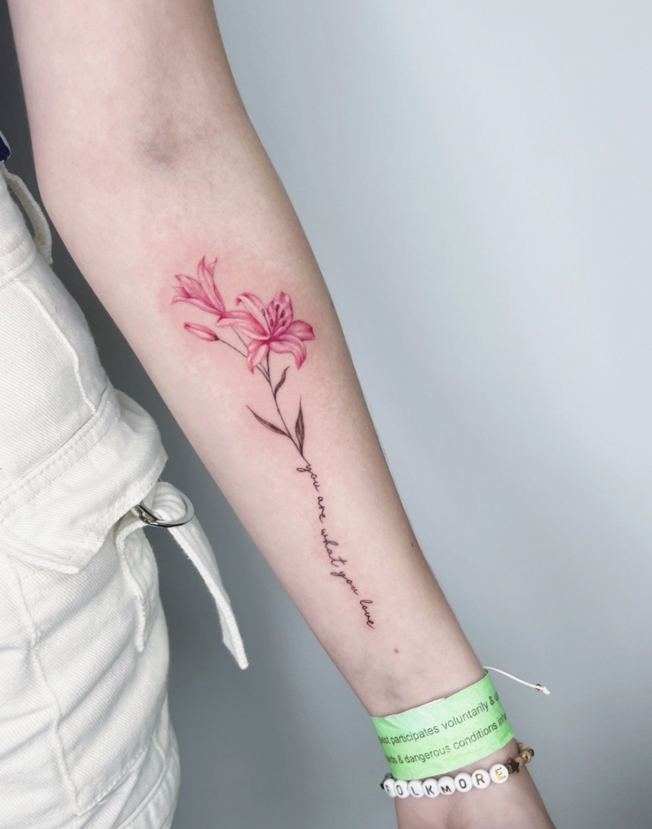 A person's arm with a pink flower tattoo, a green wristband, and a bracelet with white letter beads spelling 'ILKMORE'.