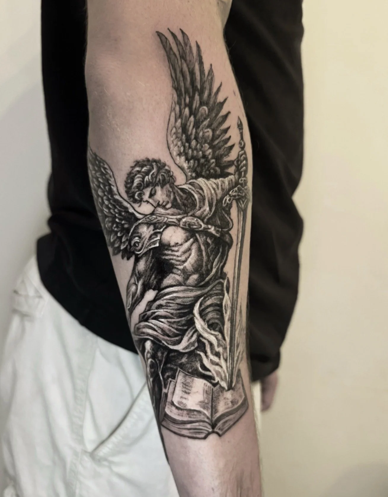 Black and gray tattoo of an angel with wings, holding a sword and a book, on a person's arm.