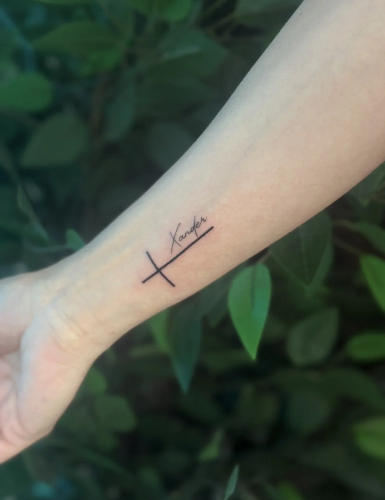 Close-up of a person's forearm with a minimalist tattoo of a horizontal line intersected by a vertical line, and the word 'Xander' written above it.