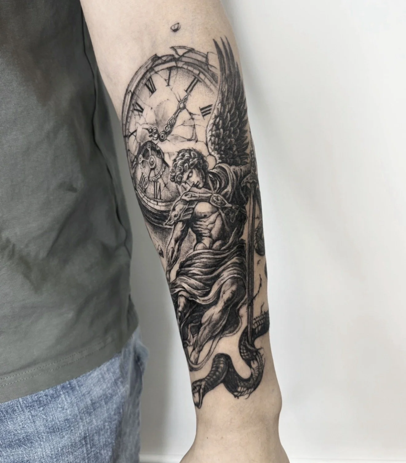 A black and gray tattoo on a person's arm depicting a mythological figure with wings, a helmet, and a cloak, holding a staff or spear, alongside a broken clock with Roman numerals and a gear, with a snake wrapped around the lower part of the arm.