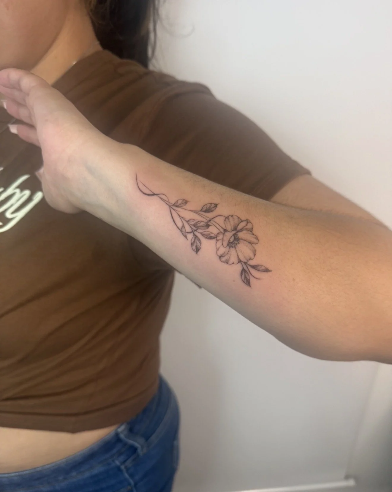 A person with a floral tattoo on their forearm, wearing a brown shirt with rolled-up sleeves, and blue jeans, standing against a plain white wall.