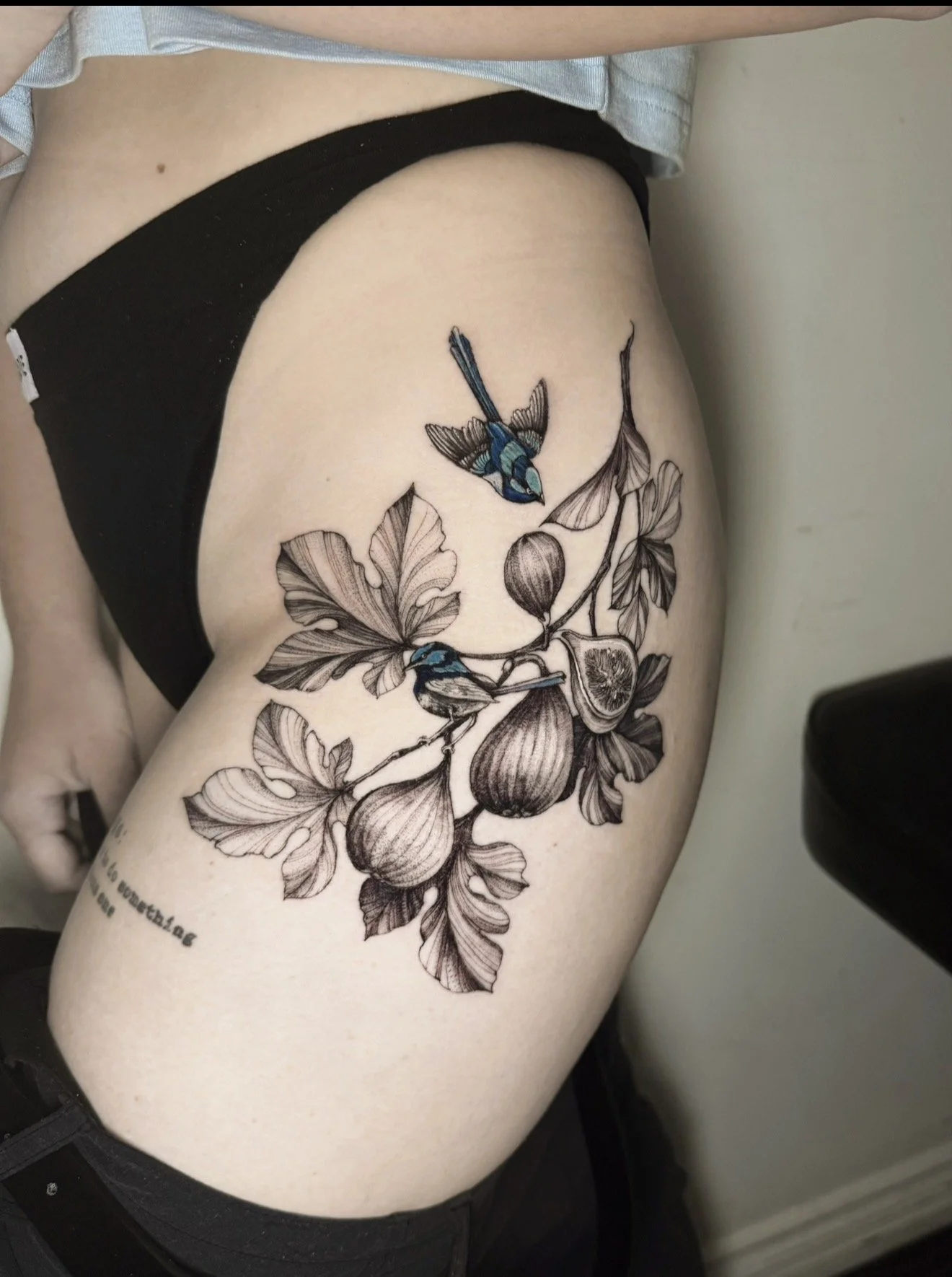 Tattoo of a branch with leaves, figs, and two colorful hummingbirds on a person's thigh.