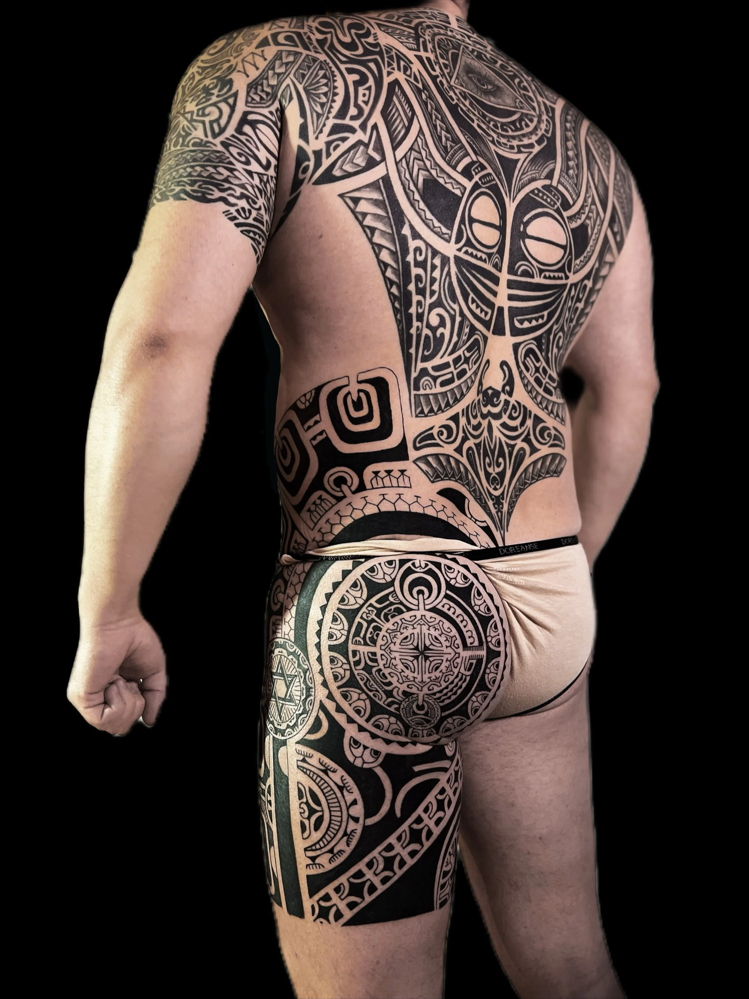 A person with extensive black tribal tattoos covering their shoulder, back, arm, and leg, featuring intricate geometric and symbolic designs on a black background.
