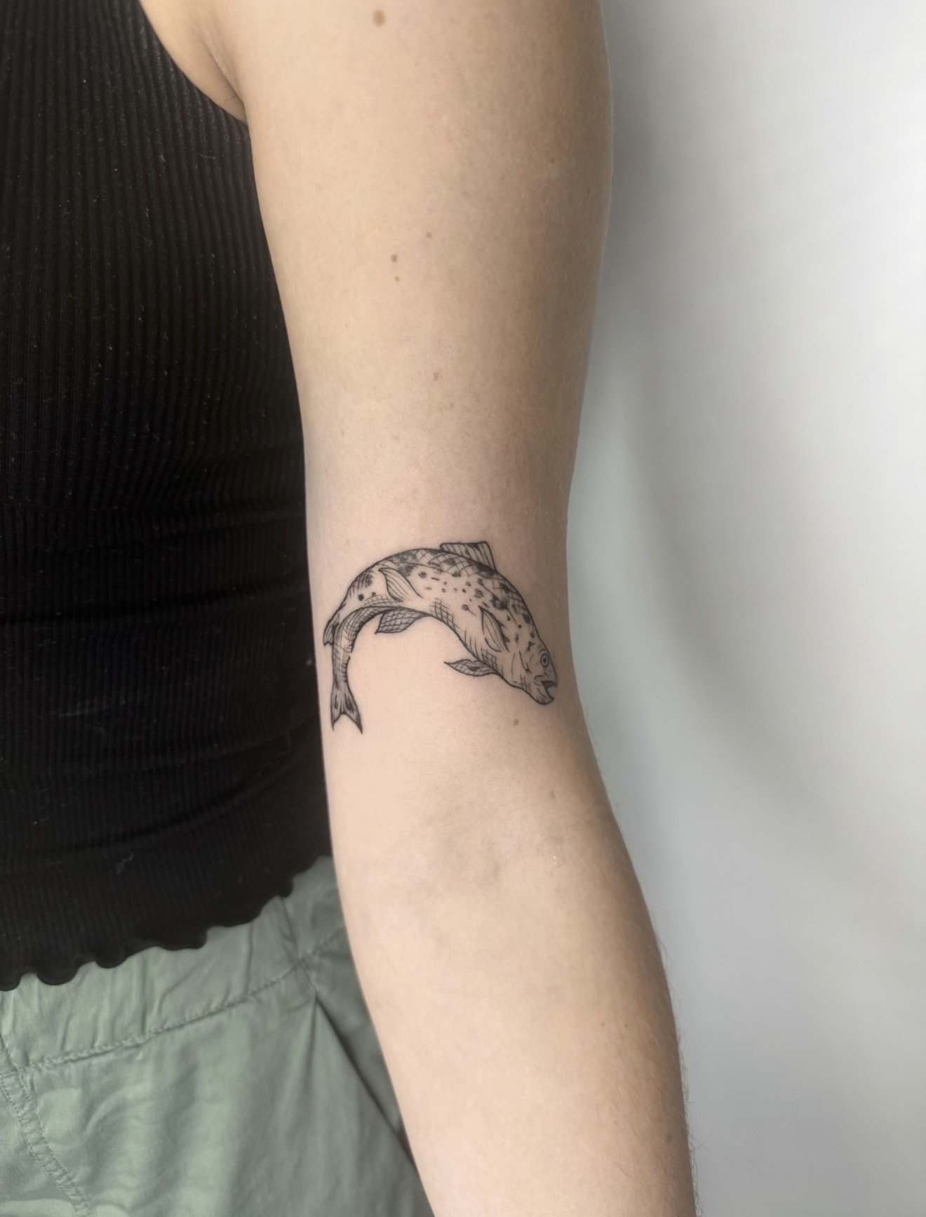 Tattoo of a fish on a person's forearm, wearing a black sleeveless top and green shorts.