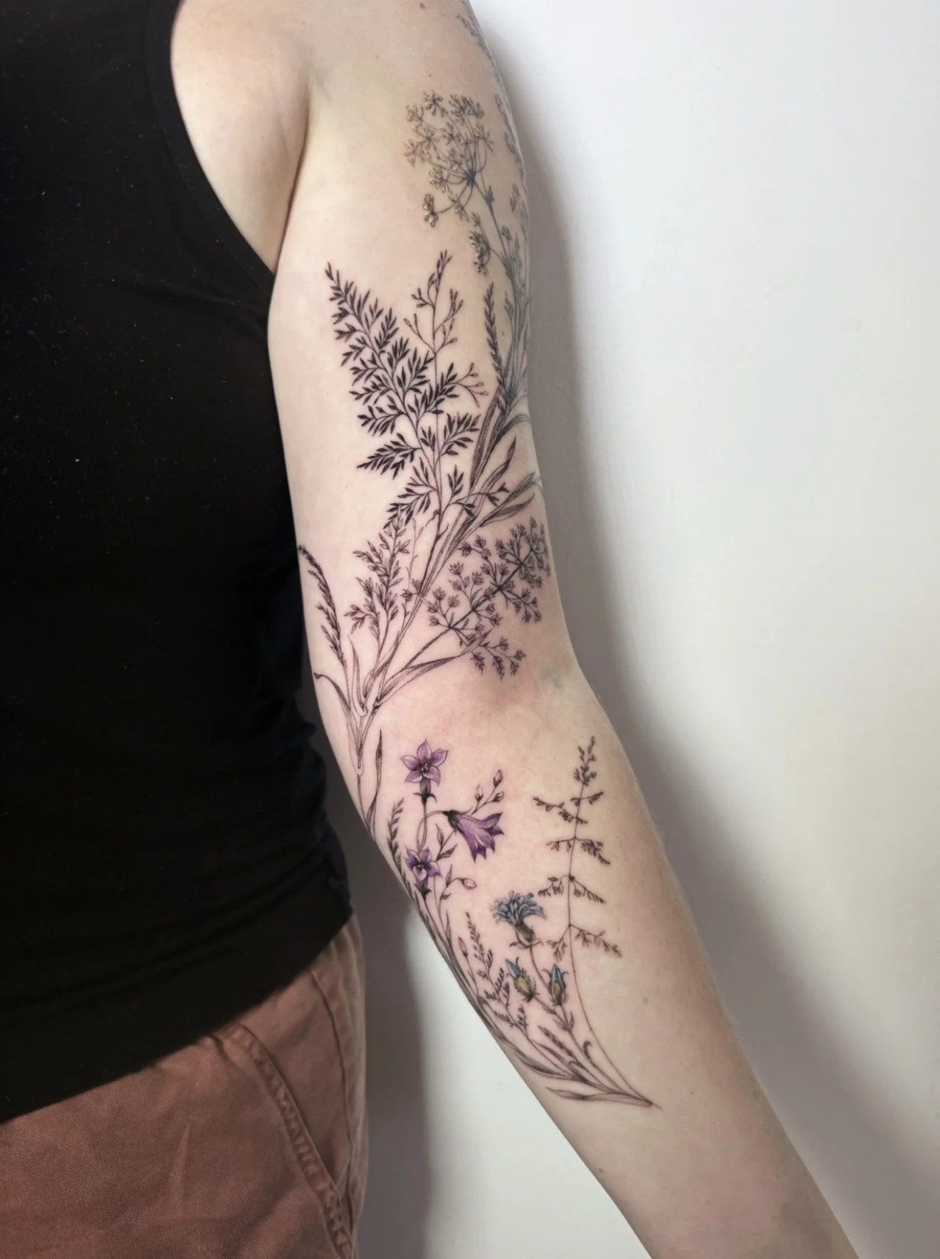 A person's arm with a detailed black ink floral tattoo featuring various types of plants and flowers.
