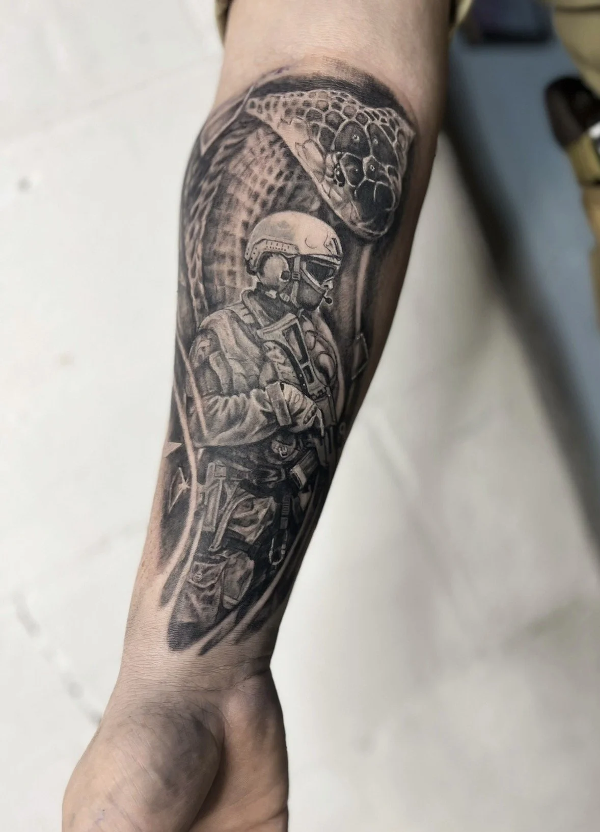 Black and gray tattoo of a soldier in tactical gear and a helmet, with a snake with a detailed head and textured scales above