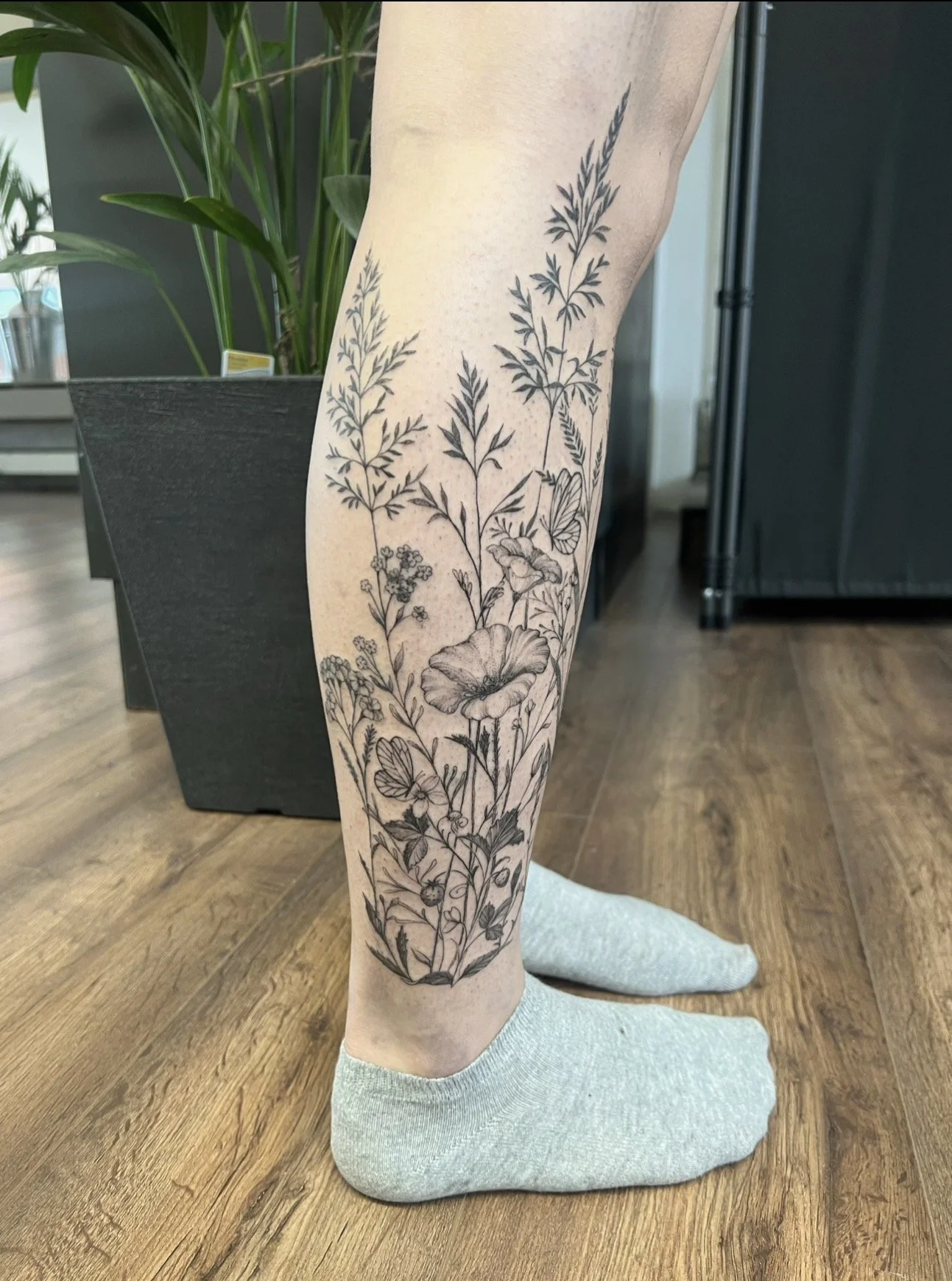 A person's leg with a detailed black and gray botanical tattoo covering from the ankle to above the knee, featuring various plants, flowers, and butterflies.