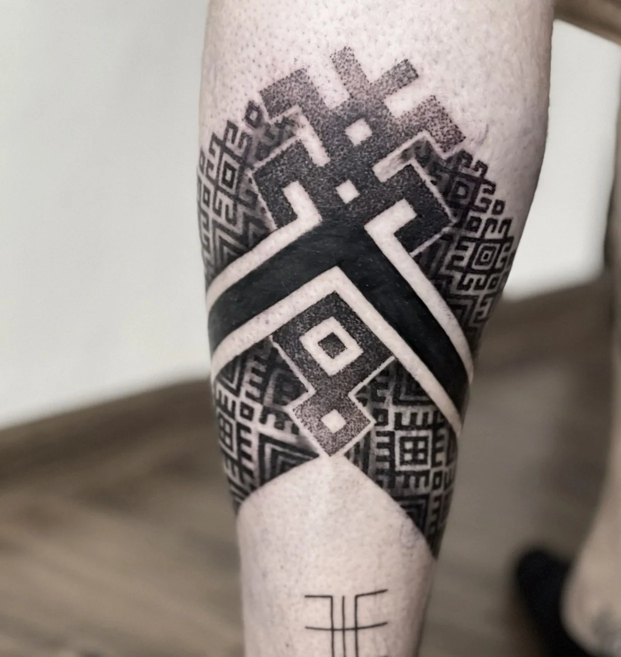 Close-up of a person's arm featuring a black geometric tattoo with intricate patterns, lines, and squares.