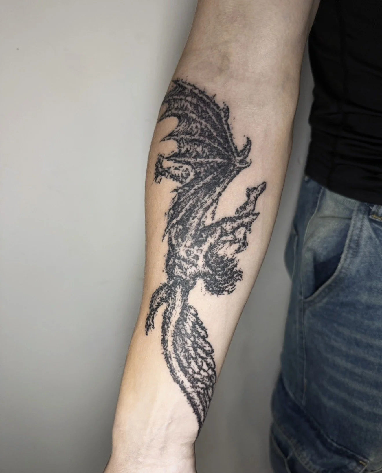 Black outline tattoo of a dragon on a person's forearm, with part of the body and wings visible, near the elbow.