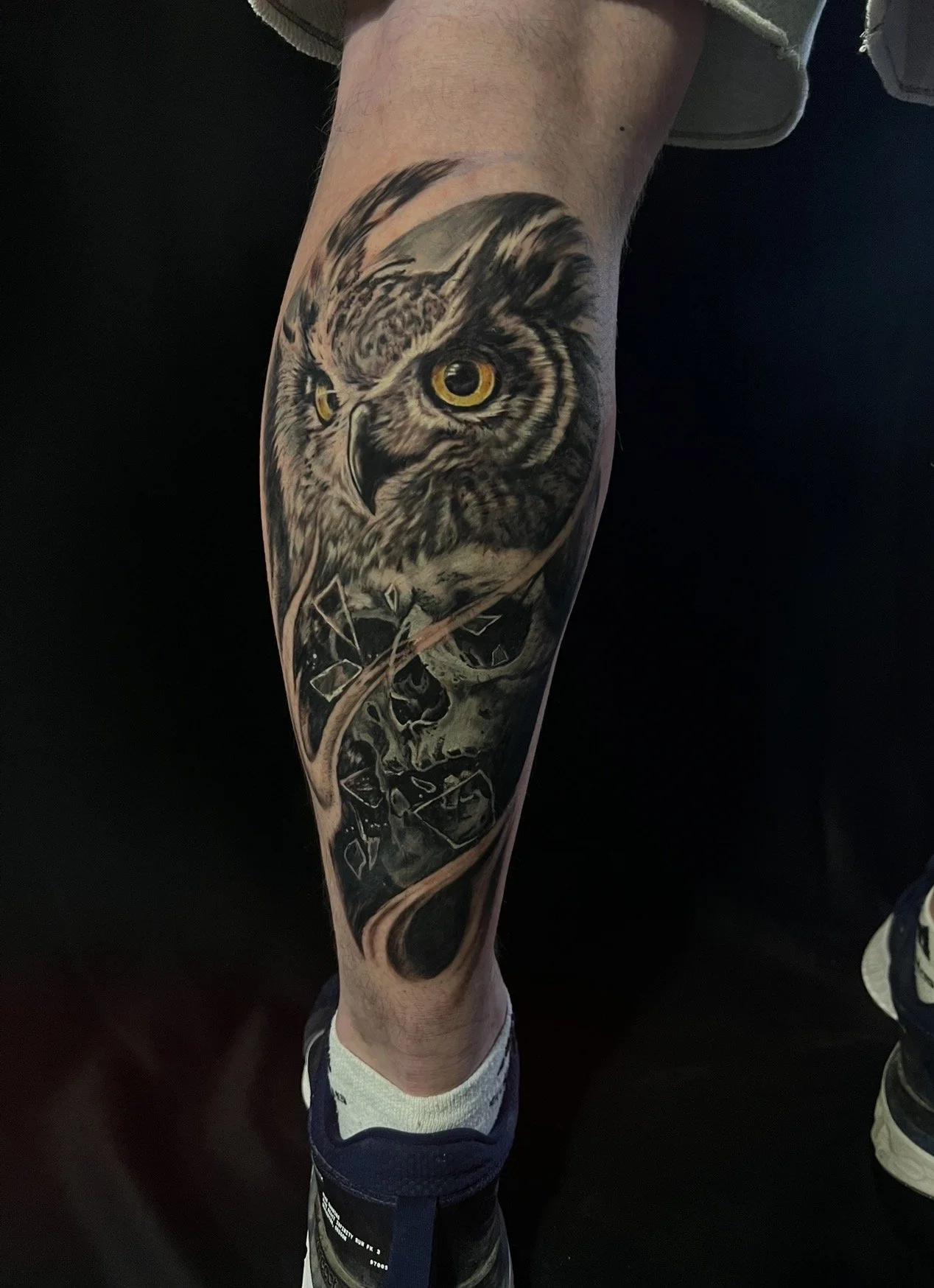 Tattoo of an owl with piercing yellow eyes on a person's leg, featuring detailed feathers and a background of falling leaves.