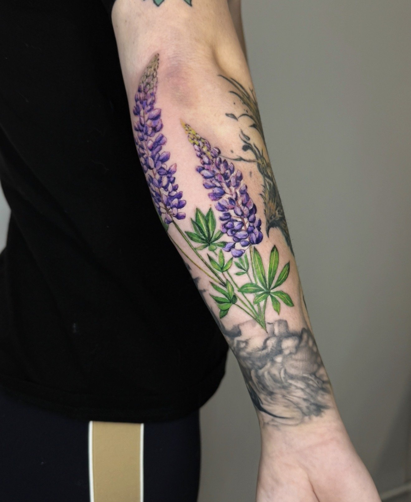 Tattoo of purple lupine flowers with green leaves on a person's forearm, with additional black and gray tattoo surrounding it.