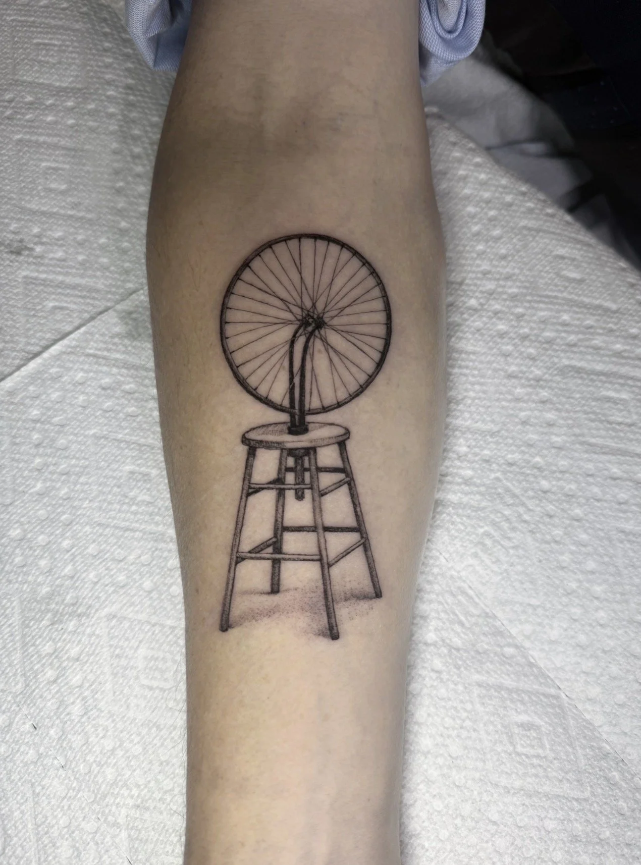 A tattoo of a vintage bicycle inside a bicycle rim, mounted on a stool with a three-legged base.