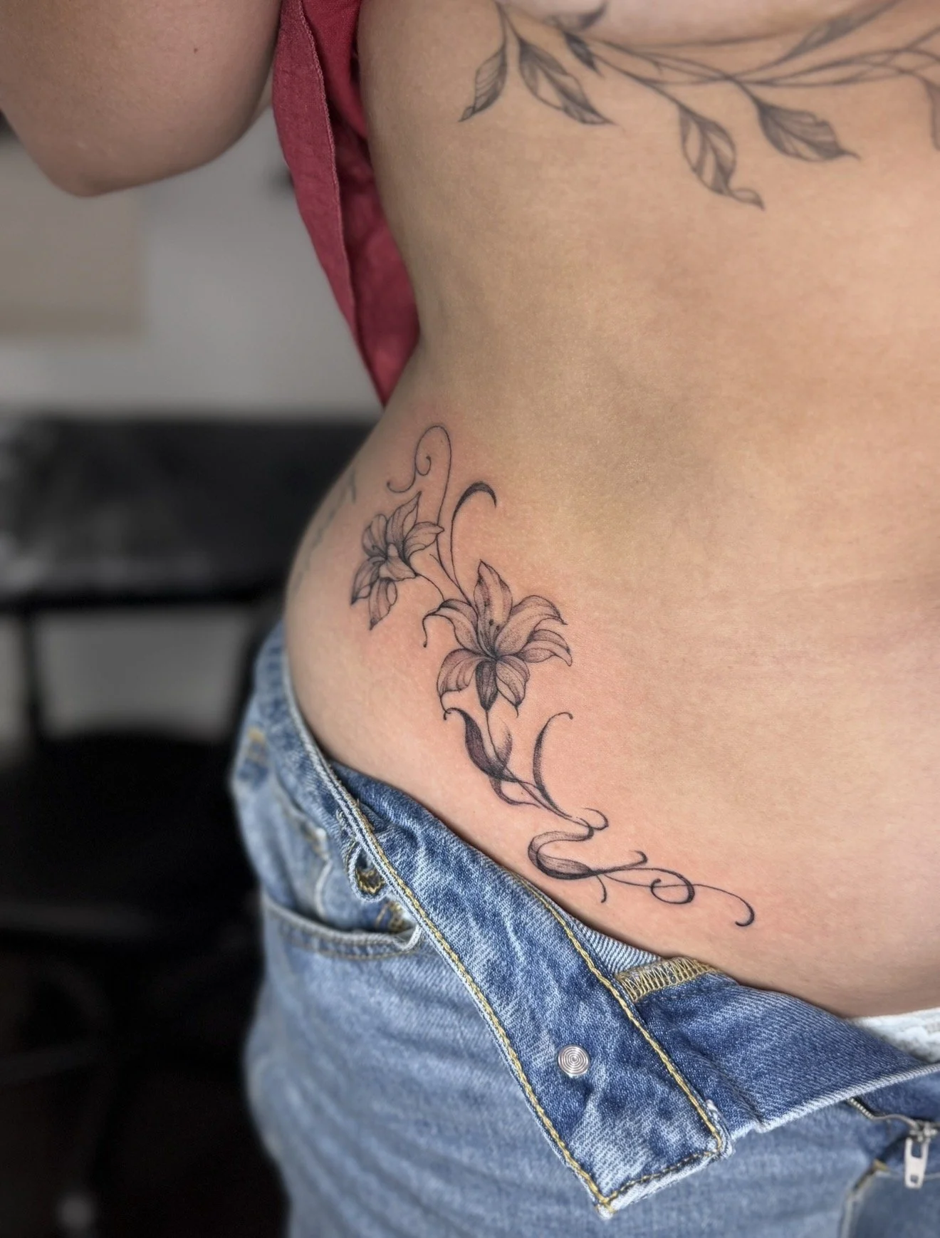 A person's side torso and waist showing a floral tattoo with lilies and leaves, along with a cursive script tattoo below, while wearing blue jeans and a red top.