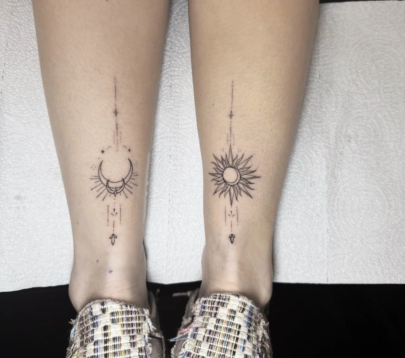Tattoo of celestial symbols on person's lower legs, featuring a crescent moon and stars on the left leg, and a sun with rays on the right leg, with dotted lines and small arrows connecting the designs.