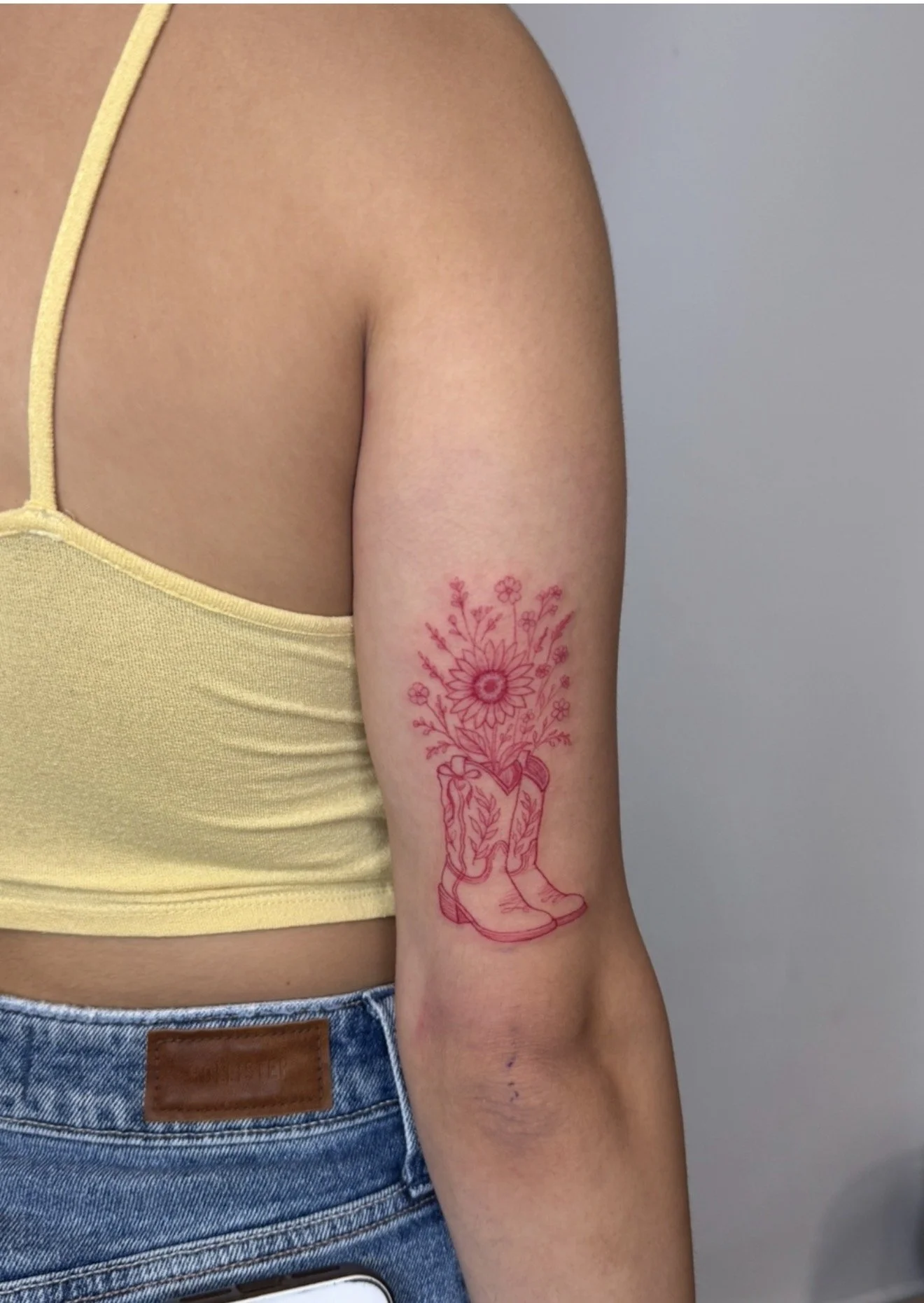 A person's upper arm with a pink line drawing tattoo of a pair of cowboy boots filled with flowers, with the arm partially covered by a yellow tank top and a pair of blue jeans.