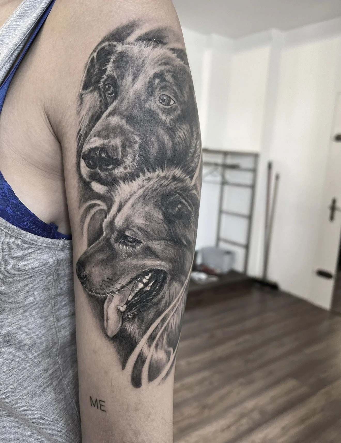 A person with a tattoo of two realistic dog portraits on their upper arm. The background shows a room with a door, a wall, and a metal shelf.