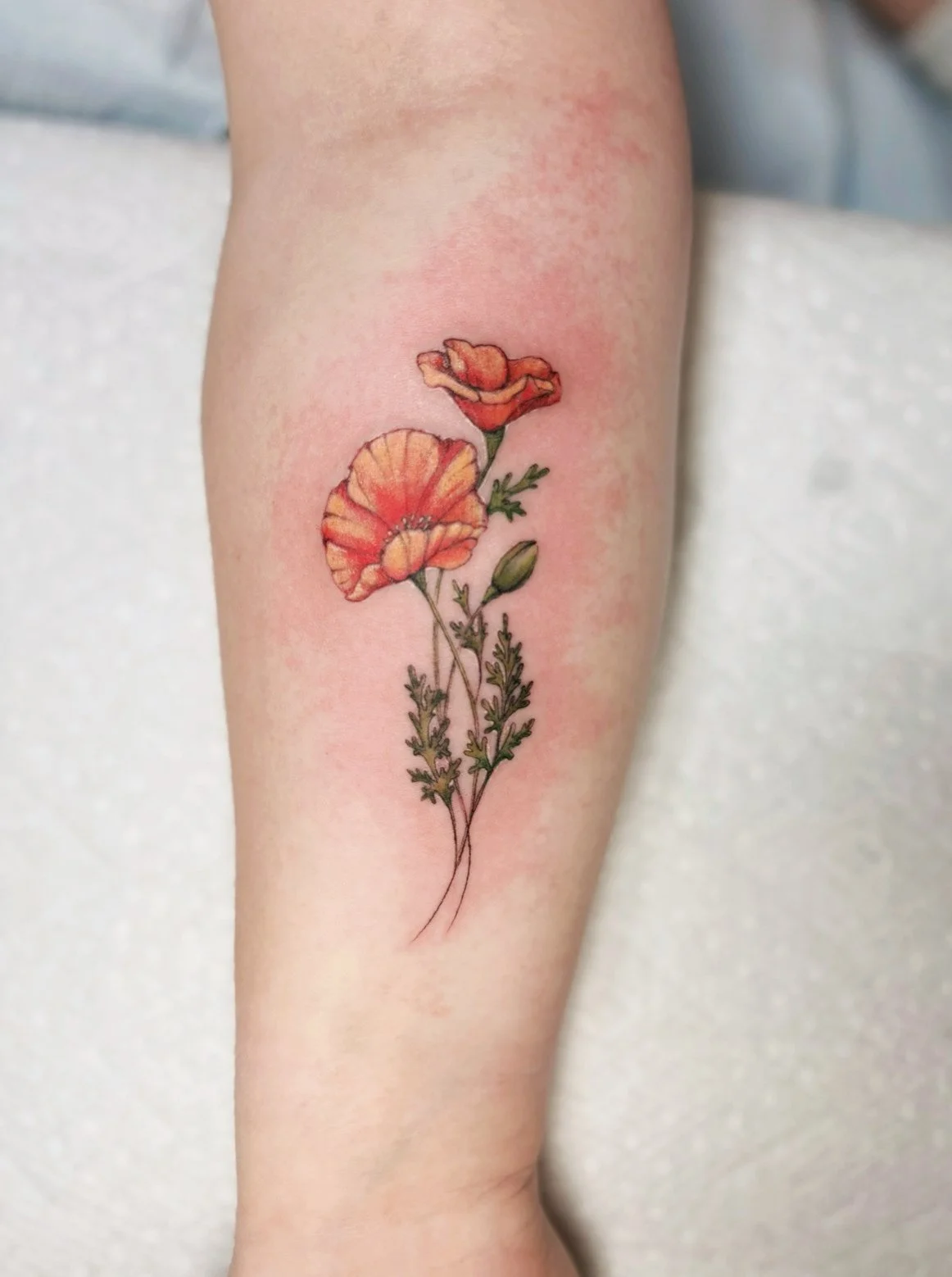 Colorful floral tattoo featuring orange and red poppy flowers with green leaves on a person's arm.