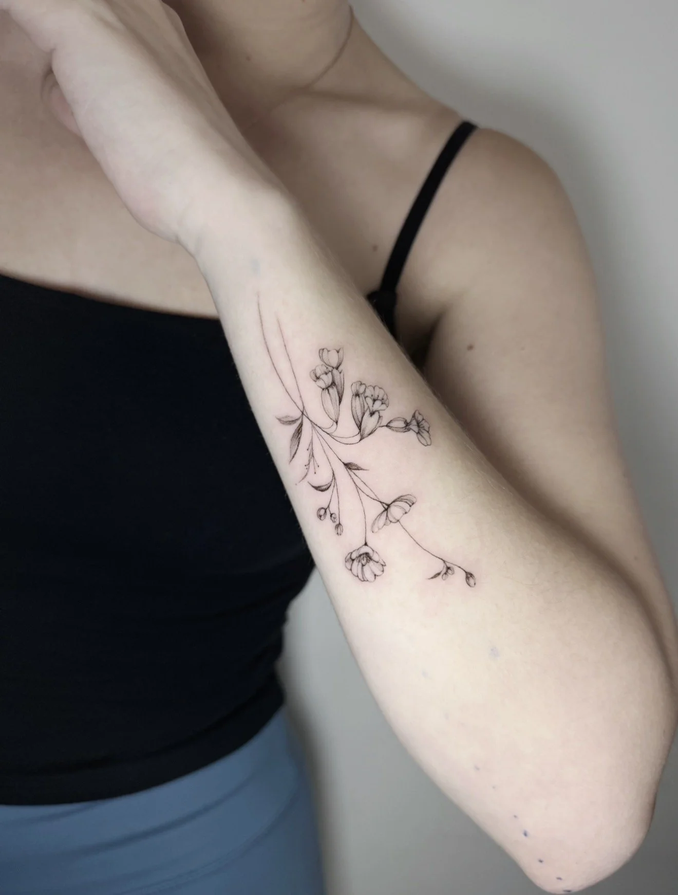 A person's arm with a delicate black outline floral tattoo on the inner forearm, wearing a black spaghetti strap top.