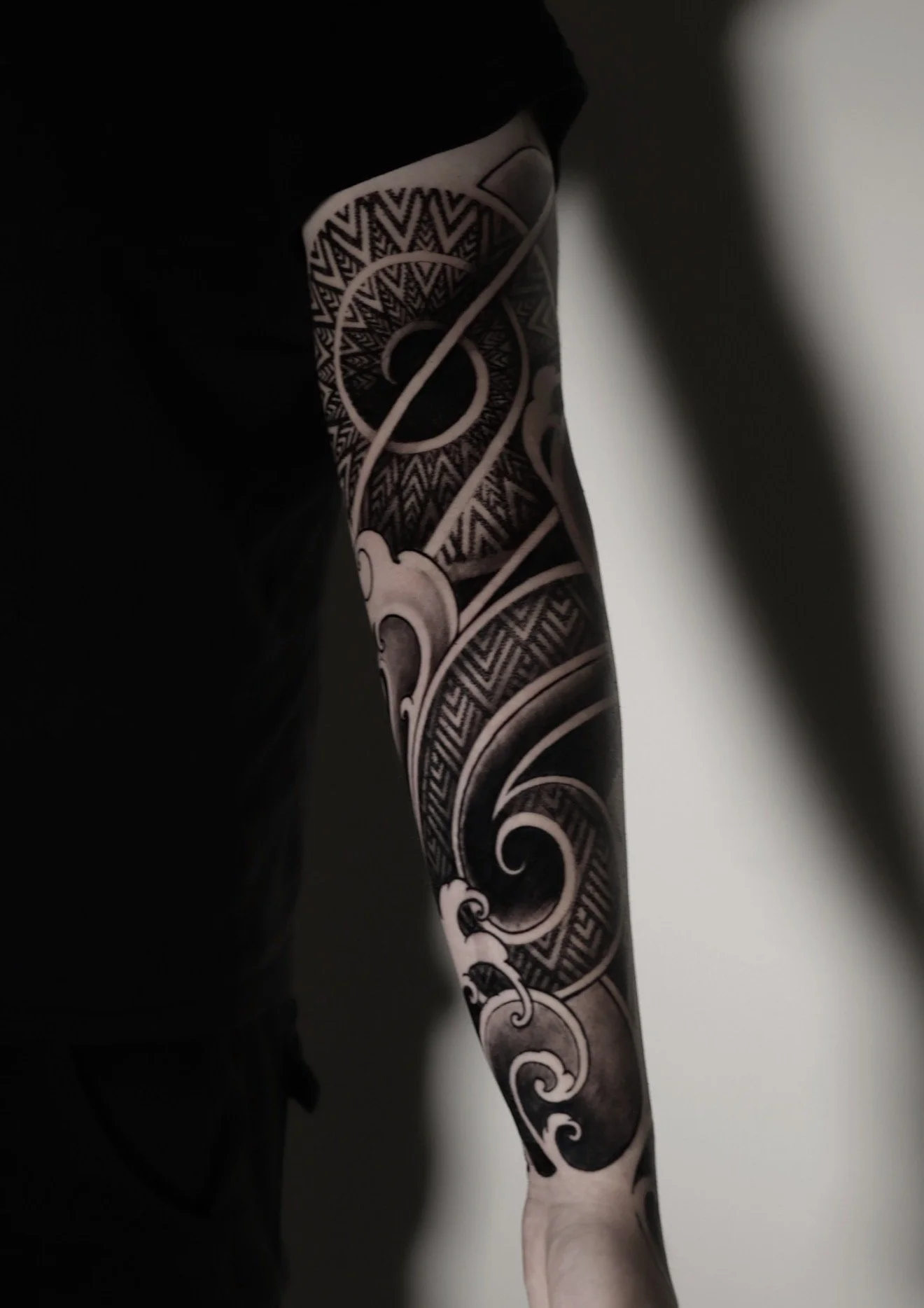 A person's arm with intricate black and white tribal-style tattoos, extending from the shoulder to the wrist, with the hand resting against a light-colored wall.