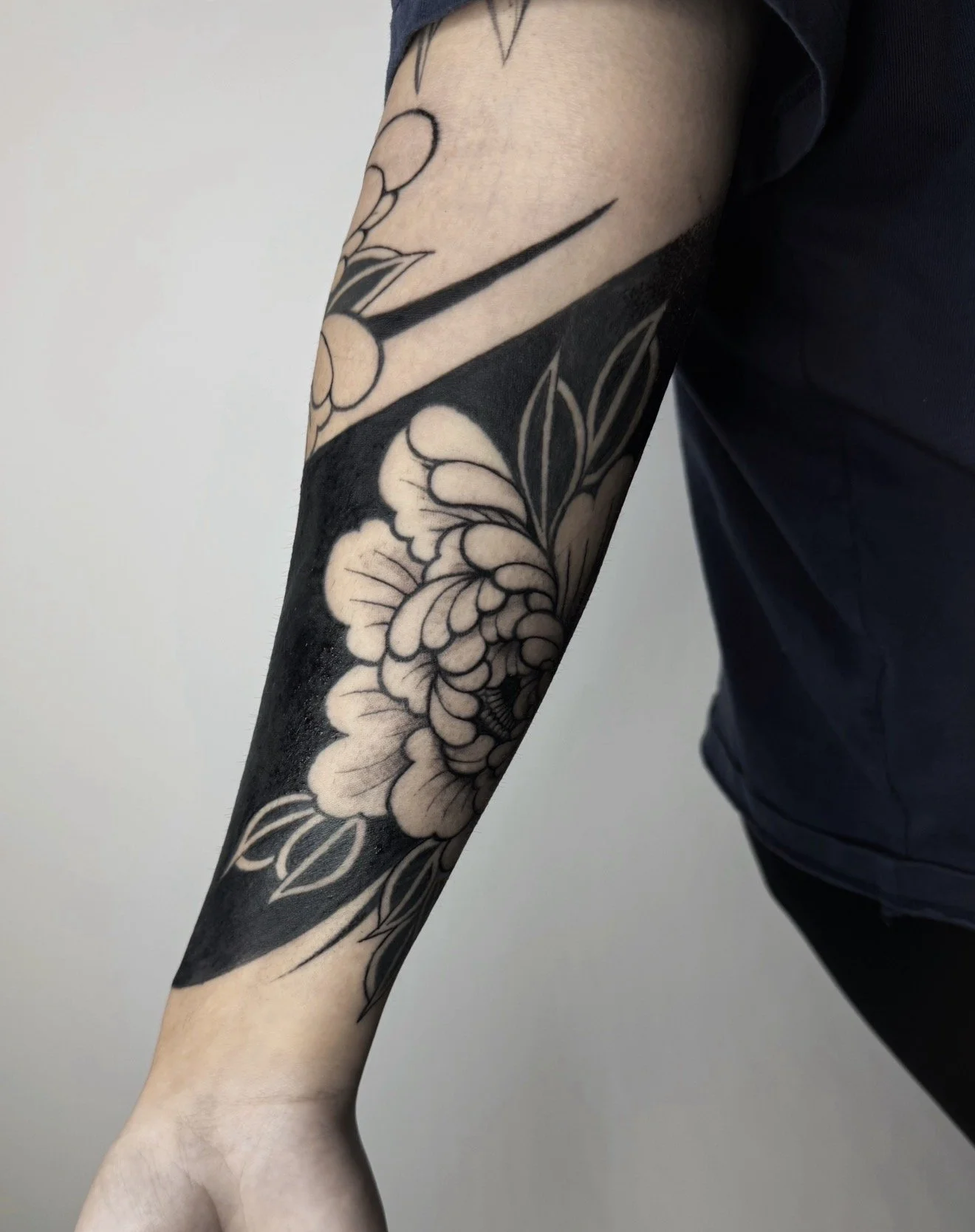 Close-up of a person's arm with a black and gray floral tattoo covering most of the forearm, with the person wearing a dark shirt and black pants.