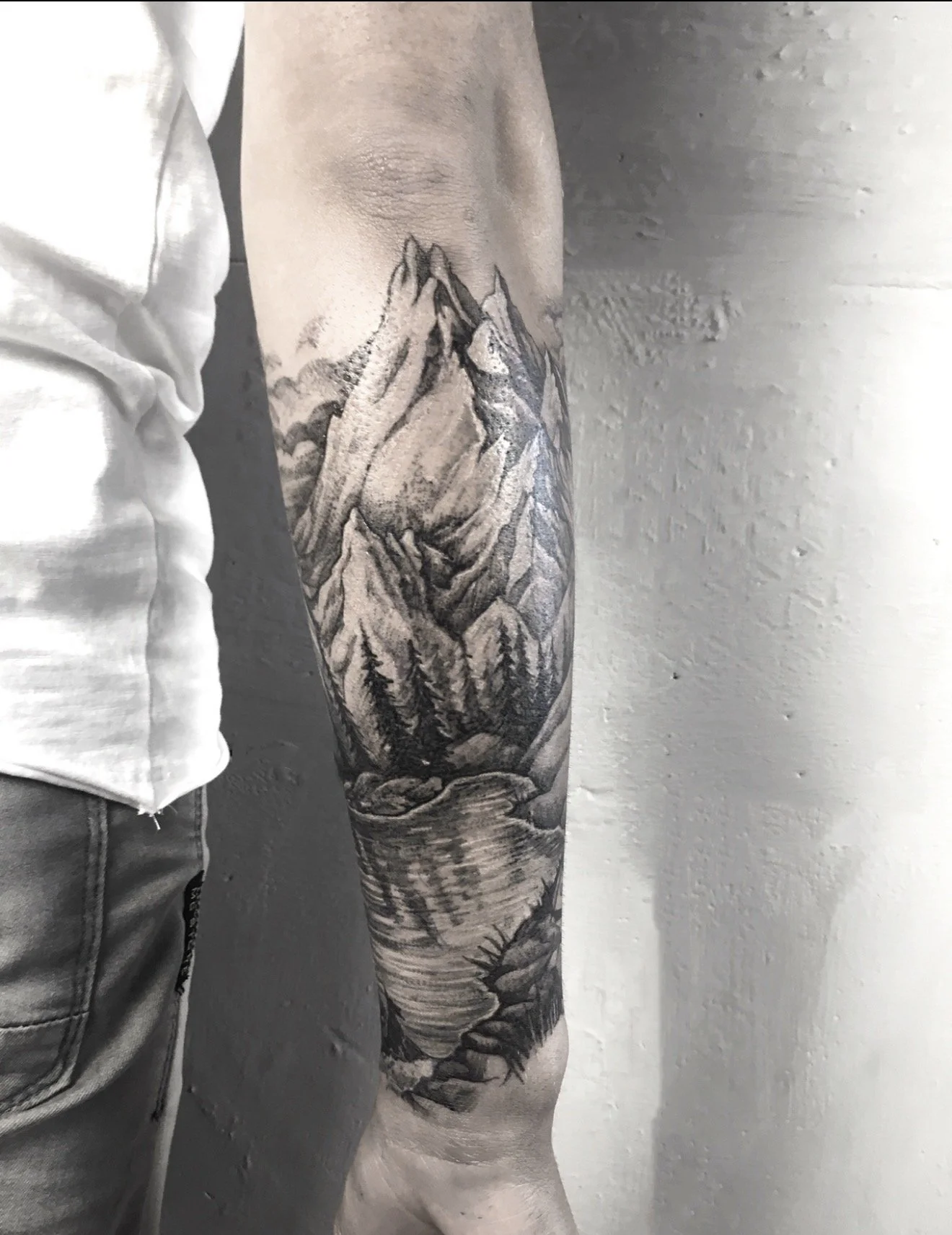 Black and gray tattoo of mountains, a river, and a forest on a person's arm, with a white t-shirt sleeve partially visible on the left.