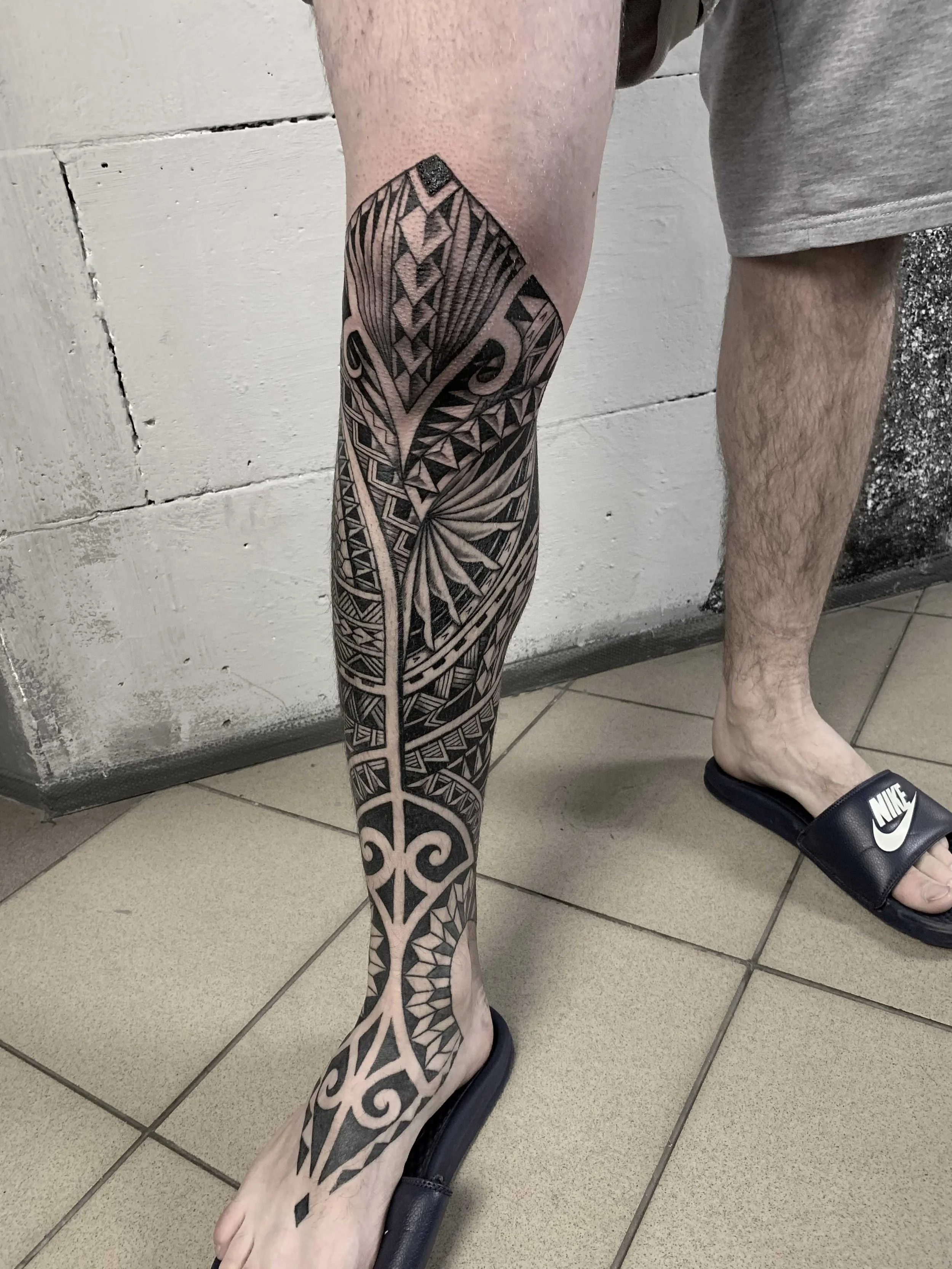 A person's leg with an extensive black ink tribal tattoo covering the thigh, knee, calf, and ankle. The tattoo features geometric and swirl patterns. The person is wearing a black Nike slide sandal on their foot, and the background includes a white w
