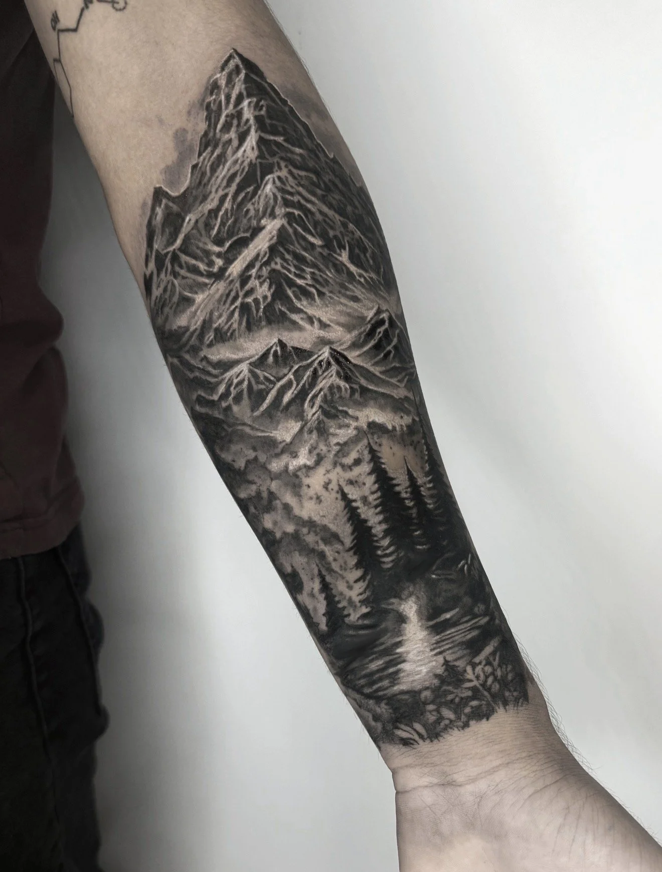 A black and gray tattoo of a mountain landscape, featuring tall mountain peaks, a lake, and trees, covering most of a person's forearm.