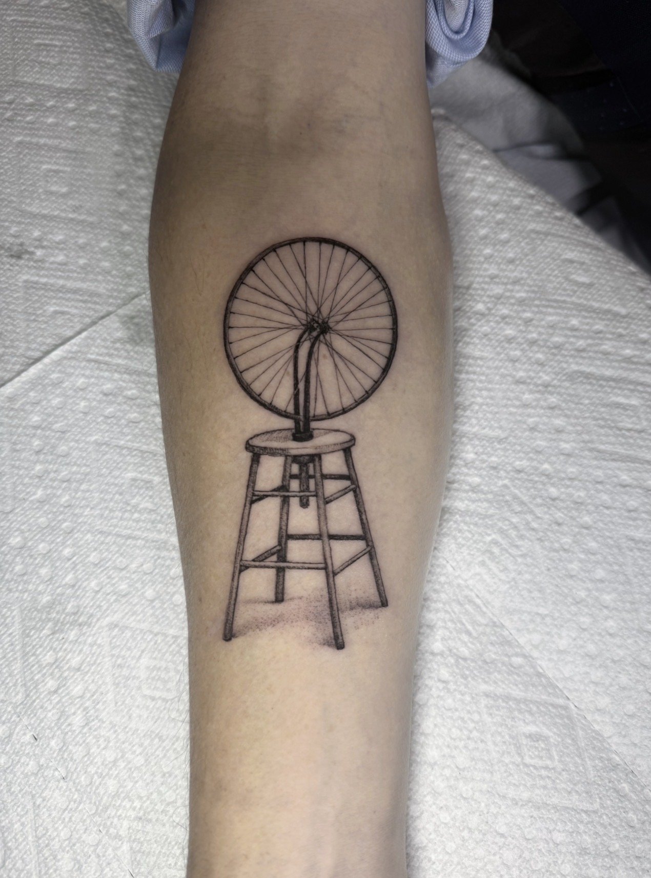 Tattoo of a bicycle wheel mounted on a stool, with the stool and wheel forming a creative design on a person's leg.
