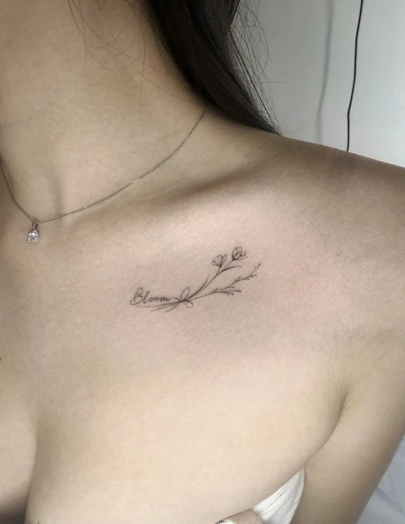 Close-up photo of a woman's upper chest and shoulder, with a delicate tattoo that says "Bloom" and has a small floral design. She is wearing a thin chain necklace with a small pendant and has dark hair partially visible.