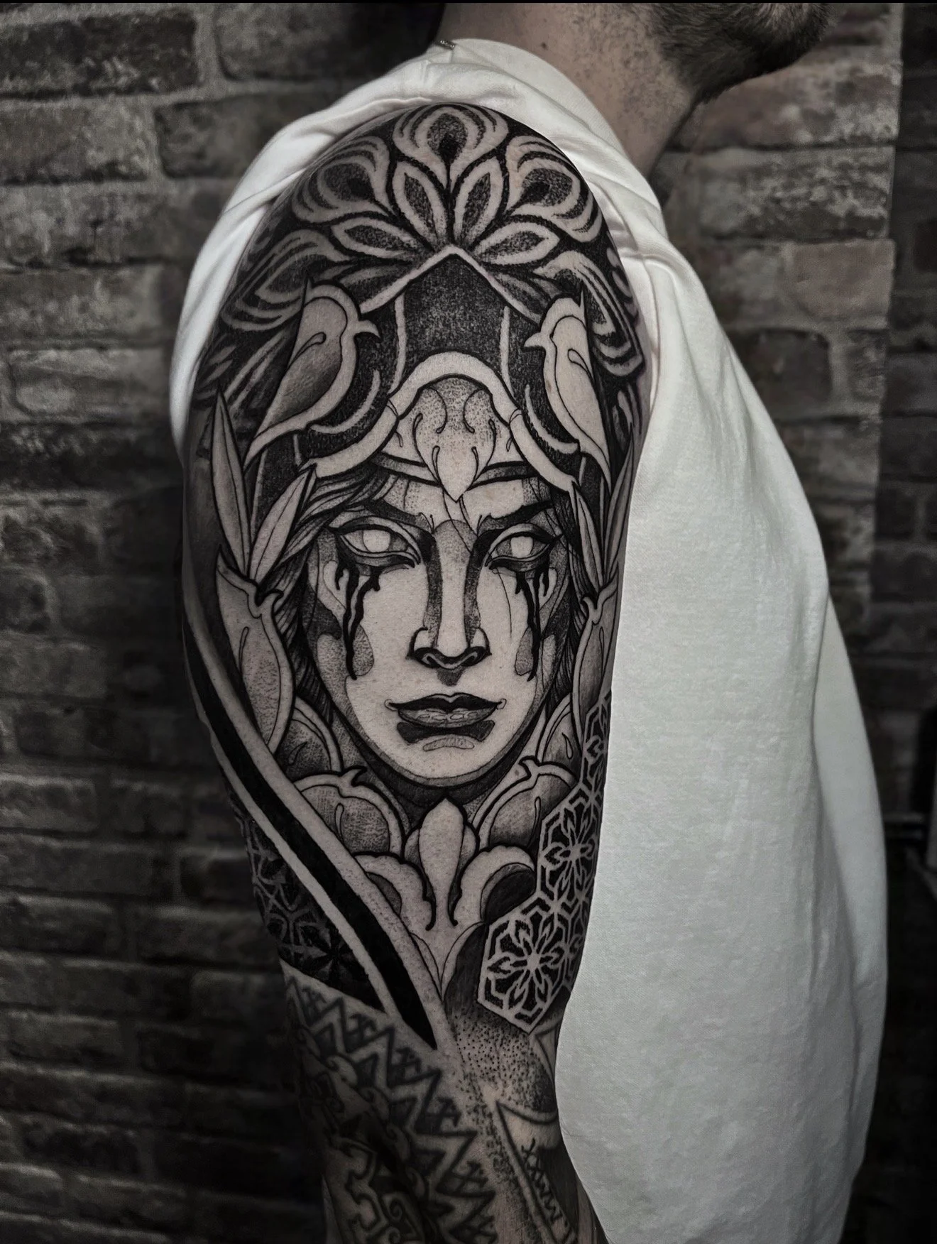 Black and gray tattoo of a woman's face surrounded by intricate patterns on the upper arm, with a brick wall background and wearing a white sleeveless shirt.