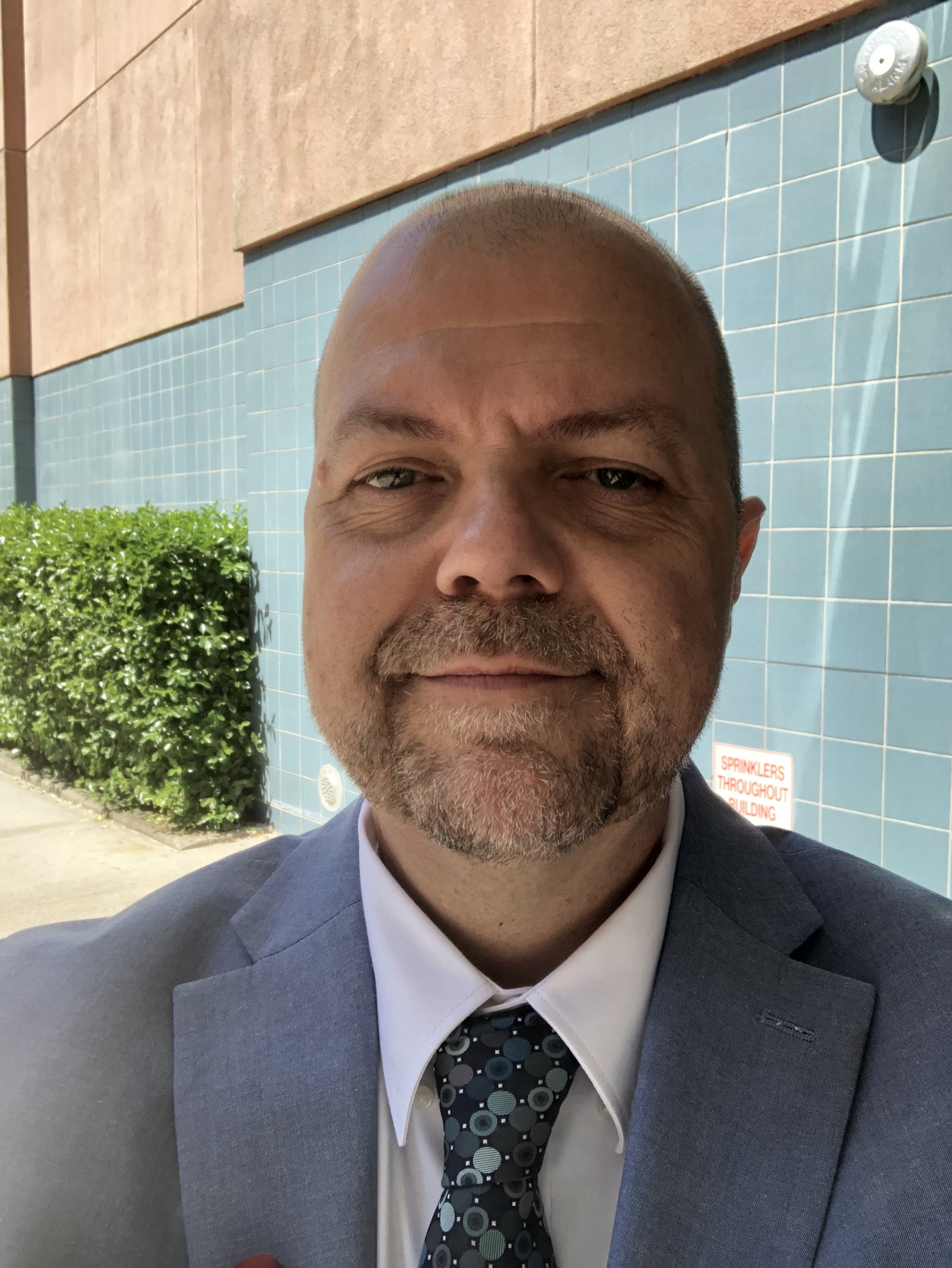 A middle-aged man with a beard and mustache wearing a purple quarter-zip sweater, smiling, standing in front of a wooden wall.