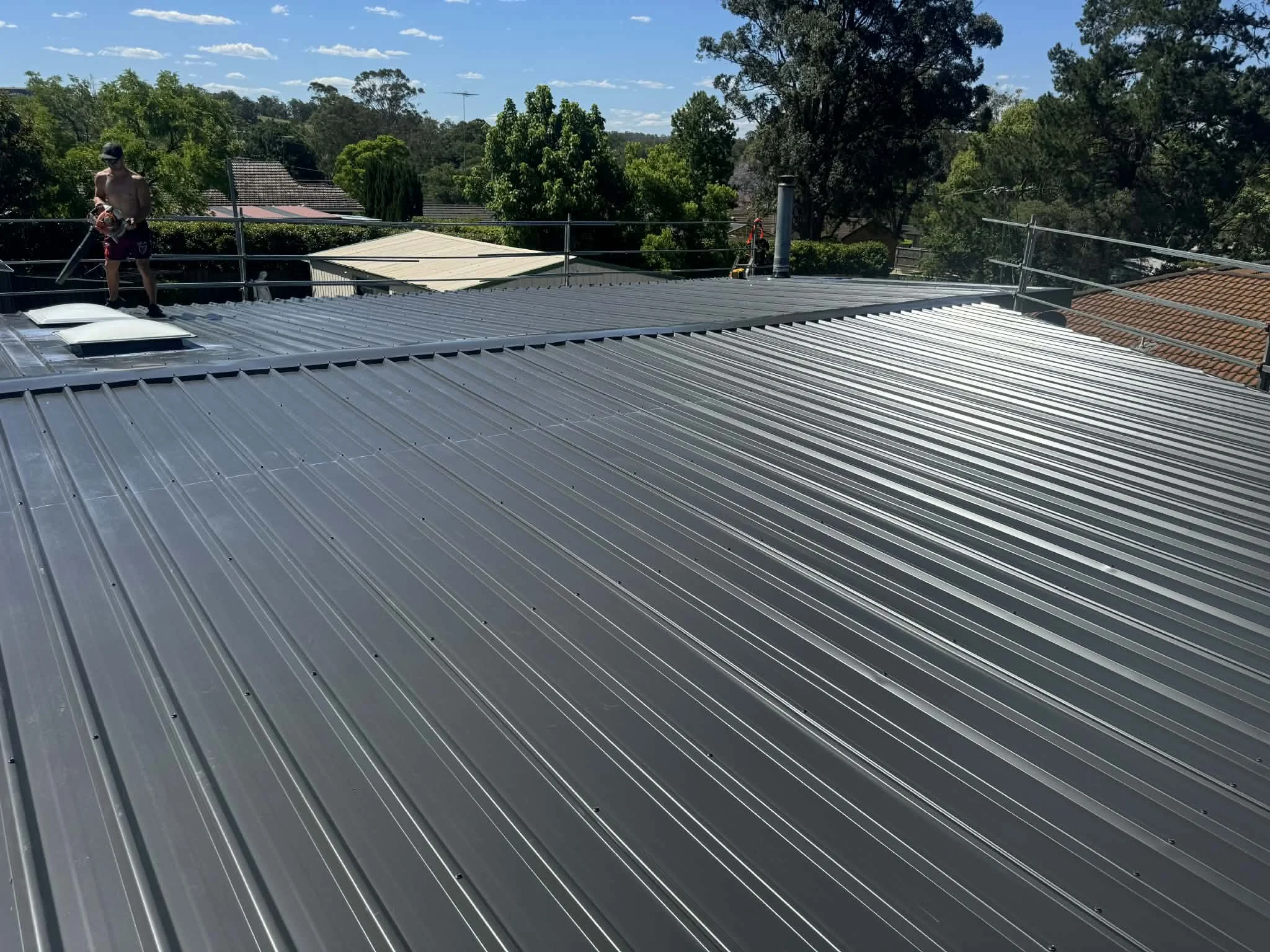 A metal roof with standing seam panels on a sunny day. Two workers are present at the edge of the roof, one holding a power tool. The background shows trees, neighboring rooftops, and a partly cloudy sky.