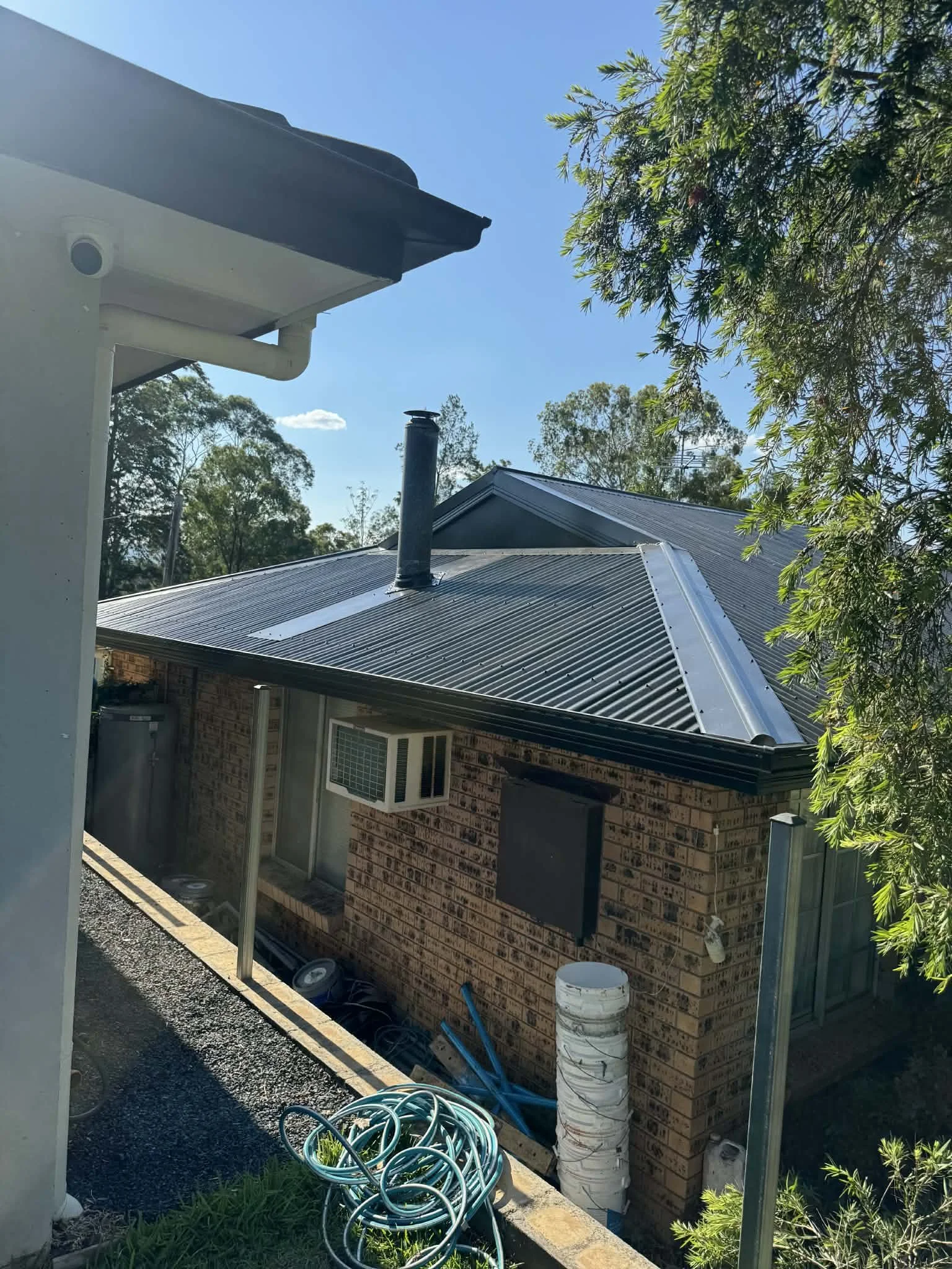 View of a house with a metal roof, a chimney, an air conditioning unit in the window, construction materials, hoses, and pipes on the ground, and a tree with green leaves.