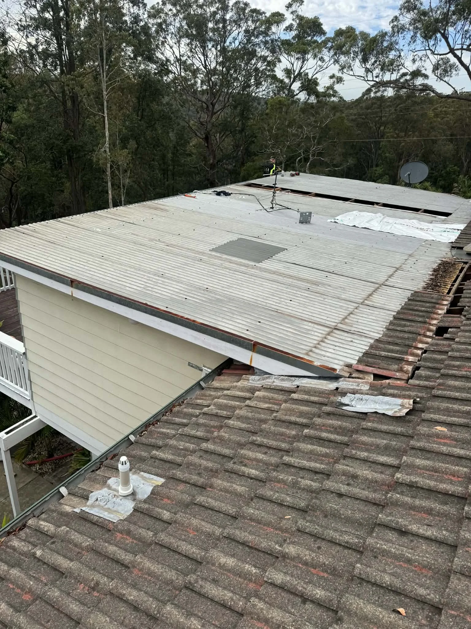 An aerial view of a house with a roof under repair. The roof has both old, weathered tiles and new metal roofing panels. Workers are visible on the metal section of the roof, and trees surround the house.