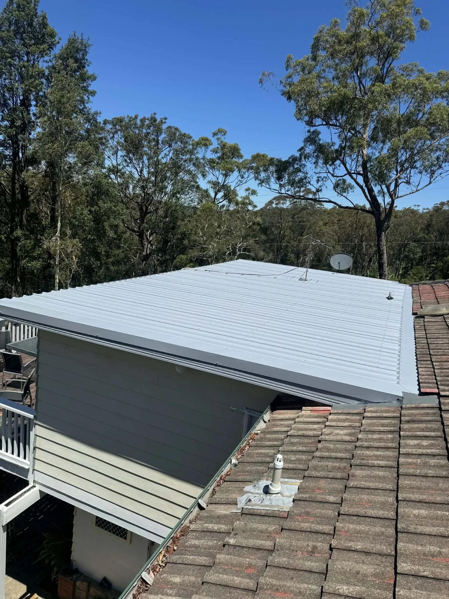 View of rooftops from a balcony showing a white metal roof, an older tiled roof, and a beige house with siding, with trees and a clear blue sky in the background.