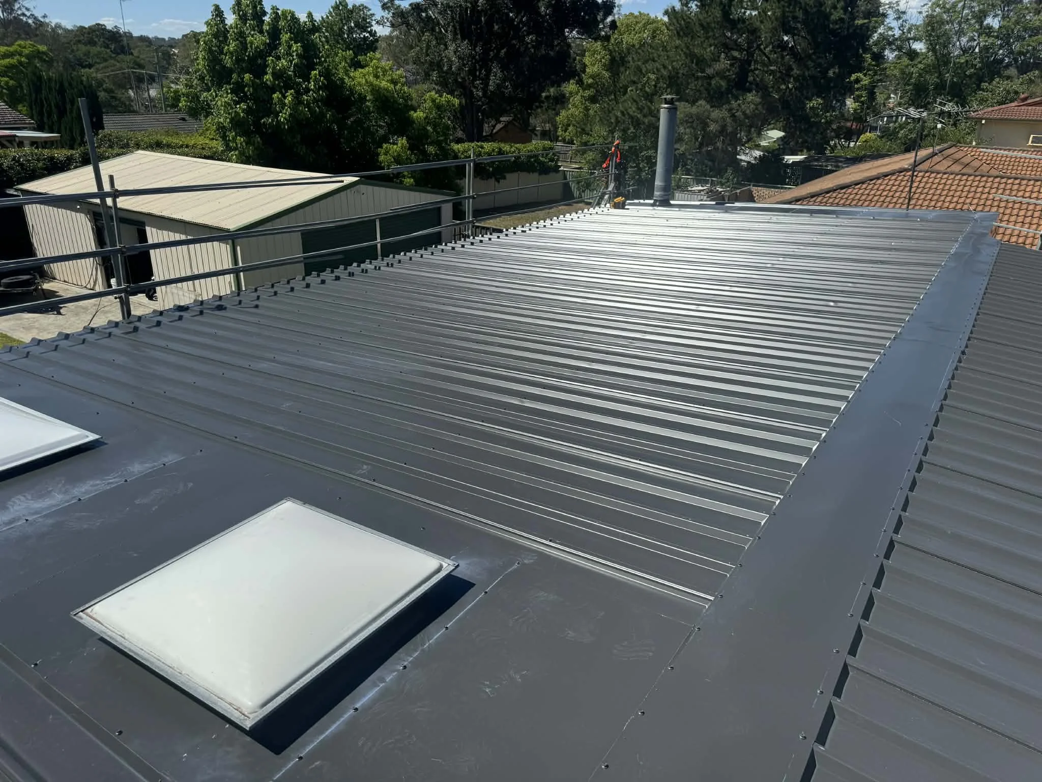 Metal roof with skylights and surrounding trees under a clear sky.