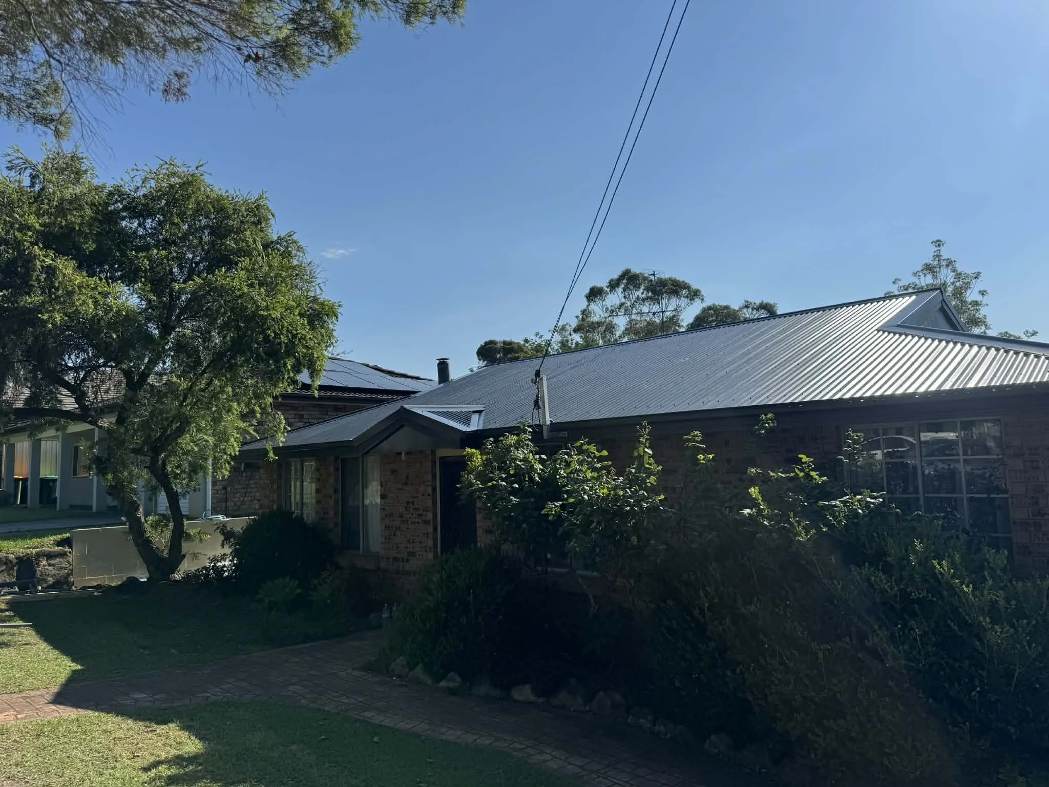 A house with a metal roof, brick walls, and arched windows, surrounded by greenery and trees in a sunny yard.