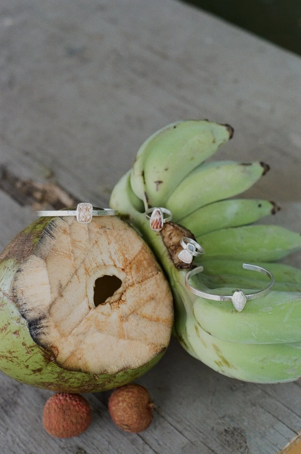 A partially cut green coconut, a bunch of green bananas, and two lychee fruits on a wooden surface, with rings placed on the coconut and bananas.
