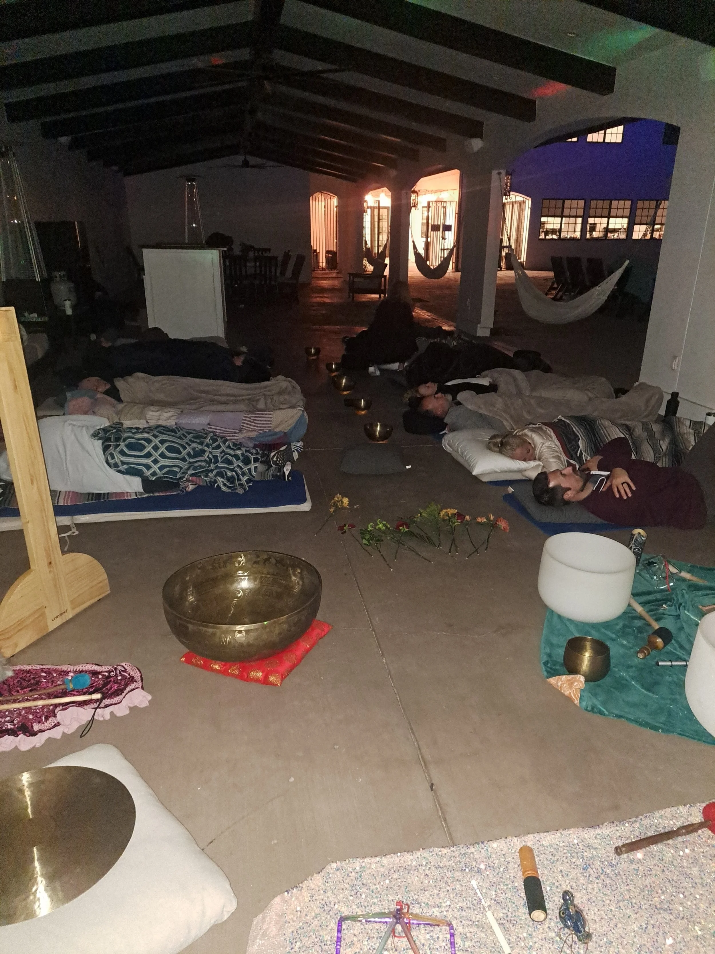 People are lying on the floor with pillows and blankets, some with bowls and instruments nearby, in a dimly lit, spacious room with hammocks and large windows showing the sunset outside.