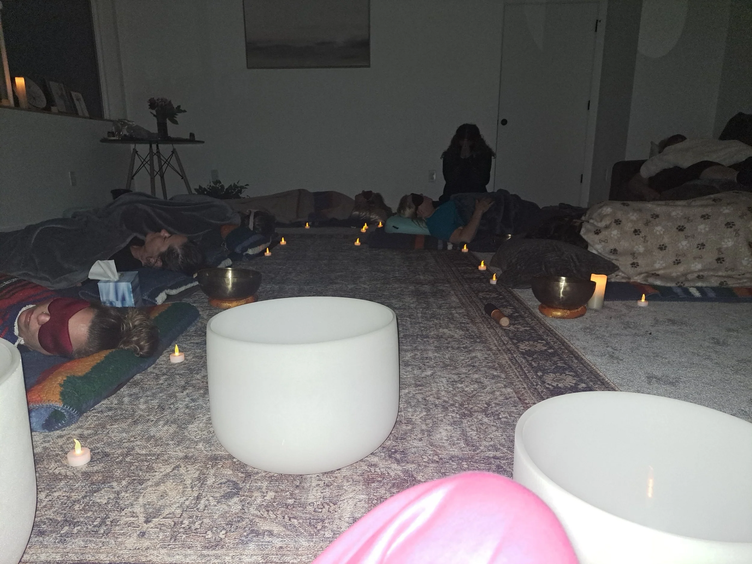 People lying on the floor with candles and singing bowls, participating in a meditation or relaxation session in a dimly lit room.