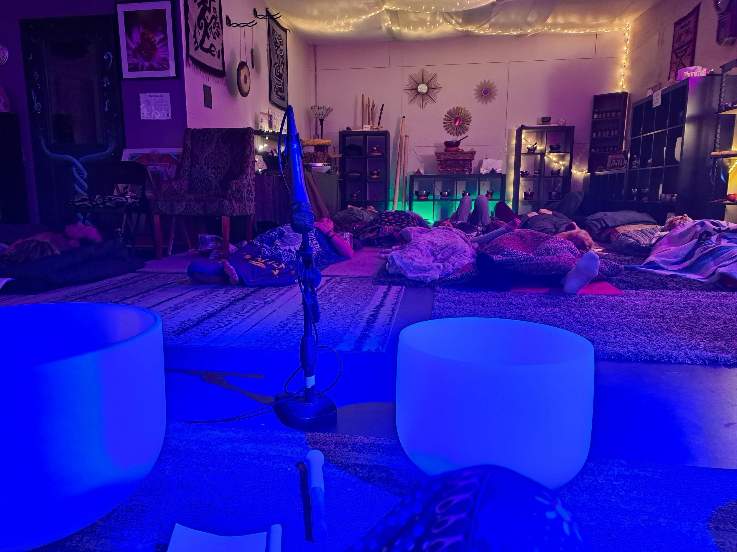 People lying on yoga mats and blankets on the floor of a dimly lit room with fairy lights, indicating a group meditation or relaxation session.