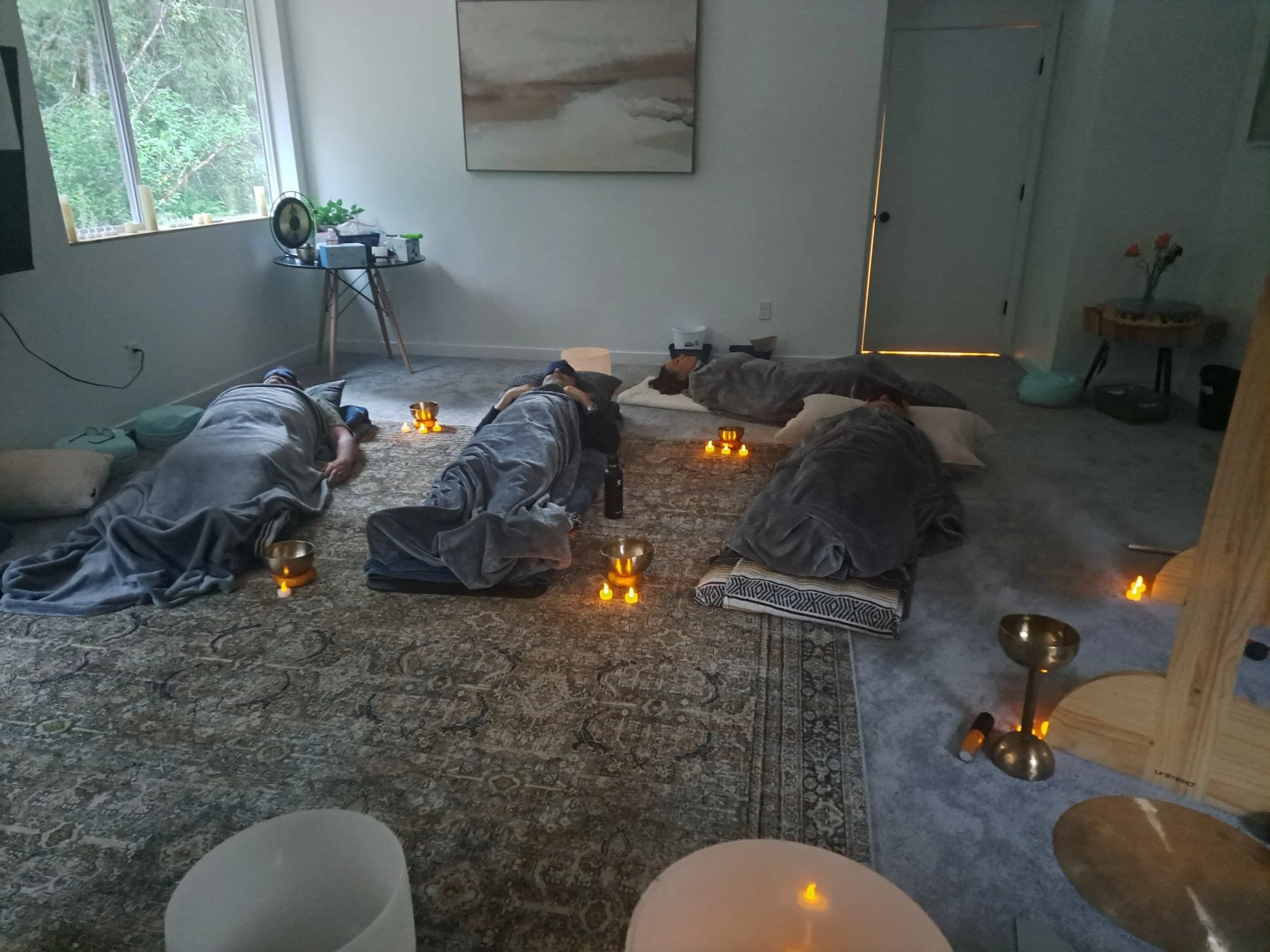 Four people lying on the floor, wrapped in blankets, participating in a meditation or relaxation session in a cozy indoor setting with soft lighting, candles, and a decorative rug.