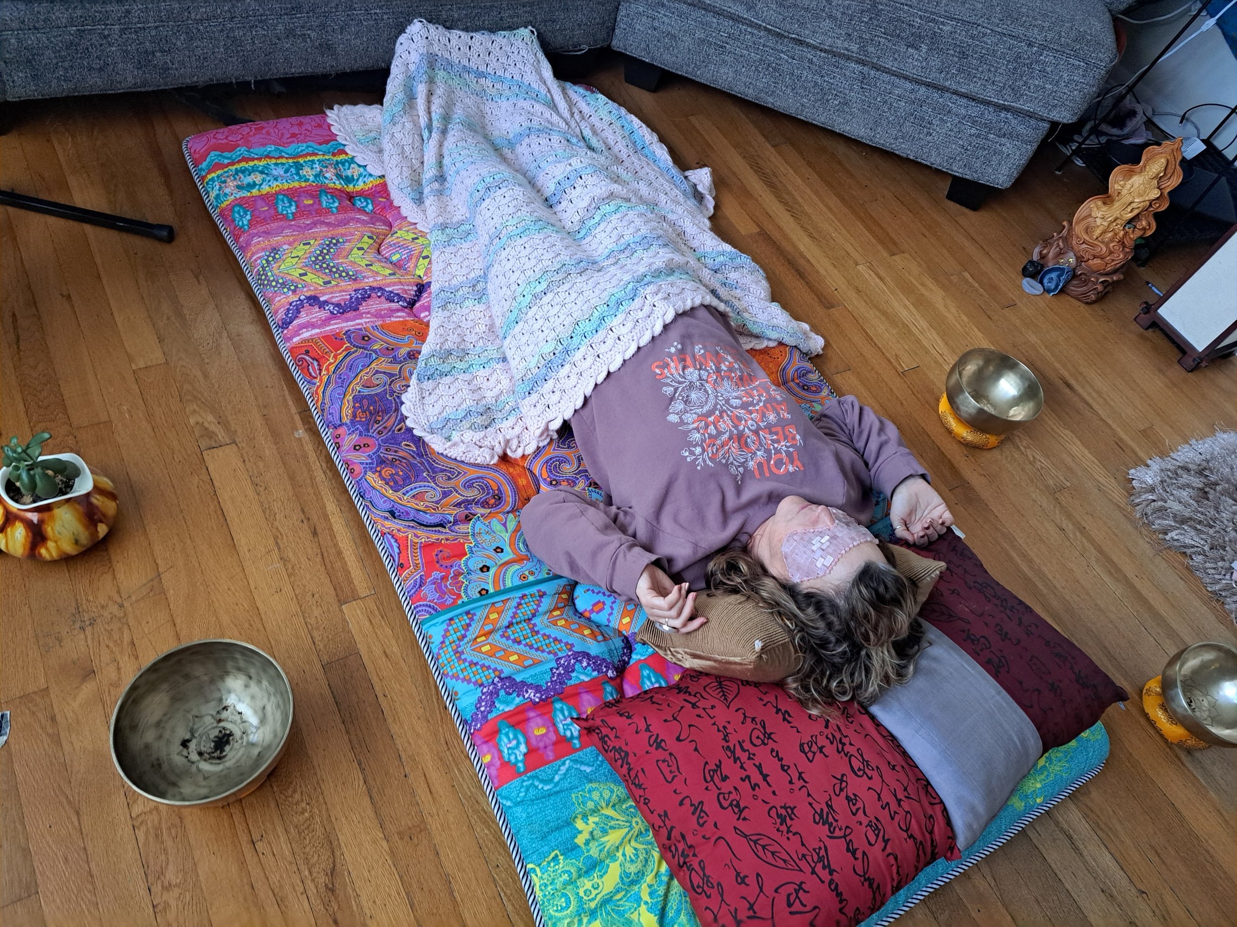 A person lying on a colorful mat on a wooden floor, covered with a light-colored blanket, with a face mask, a lavender hoodie, and pillows under their head.