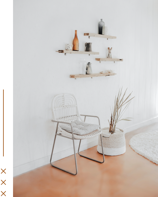 Minimalist interior with white wall, floating shelves holding decorative vases and sculptures, white wicker chair with striped cushion, tall dried grasses in a textured white planter, and a partial view of a white plush rug on a terracotta floor.