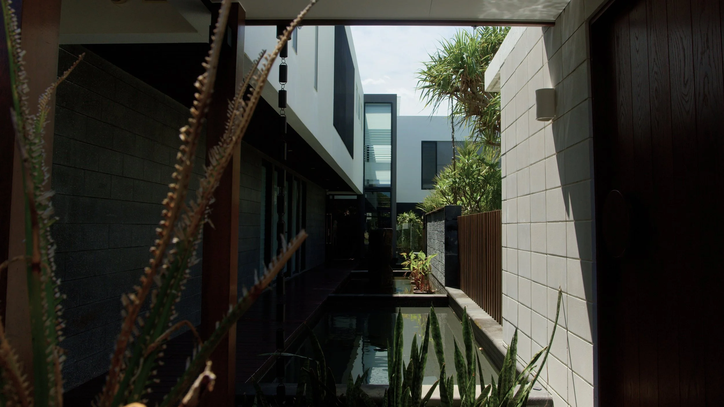 Modern house with white and black exterior, greenery, and water feature in an outdoor courtyard under a partly cloudy sky.