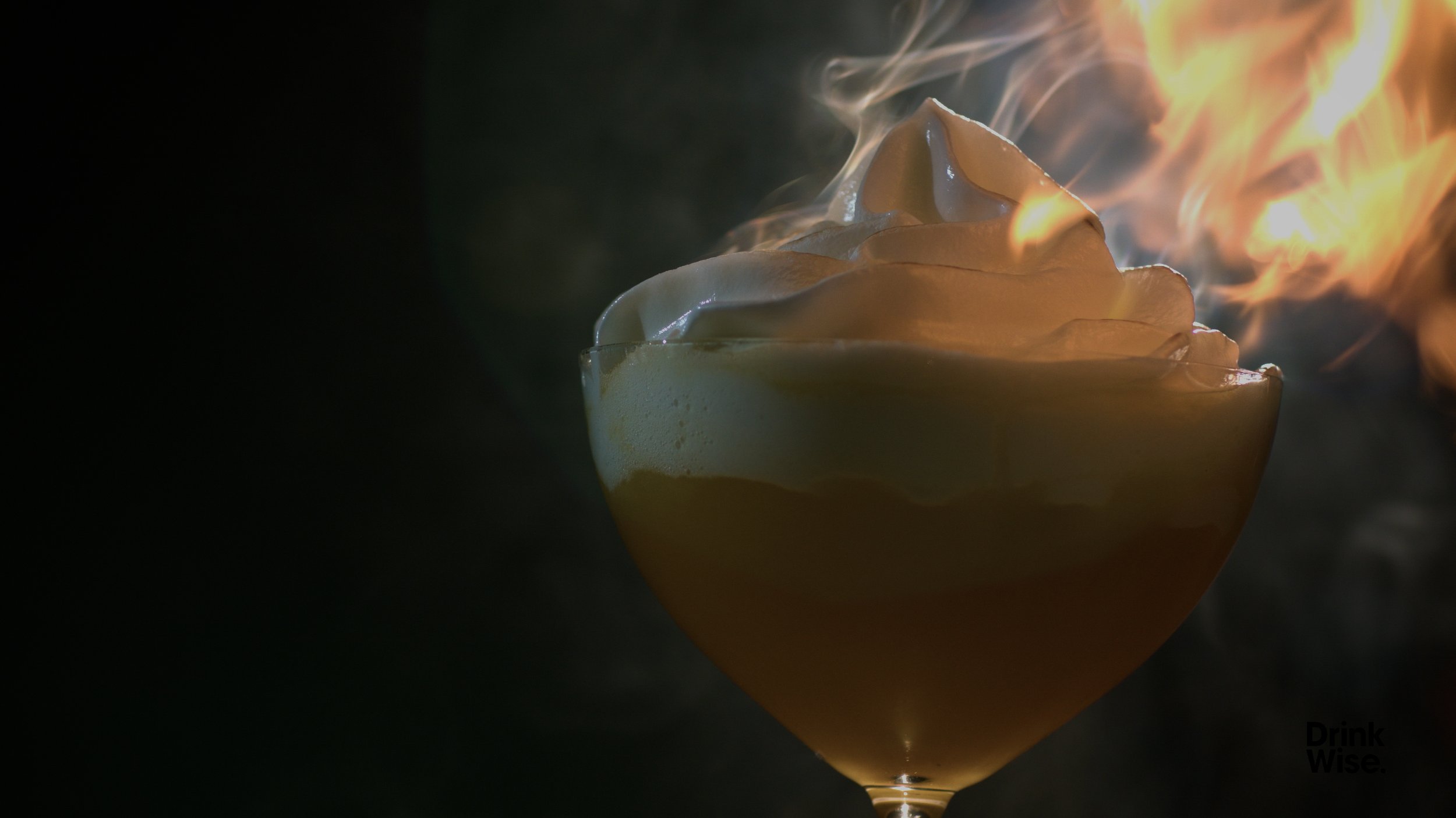 A cocktail with whipped cream on top, set in a glass, with flames and smoke rising from it.