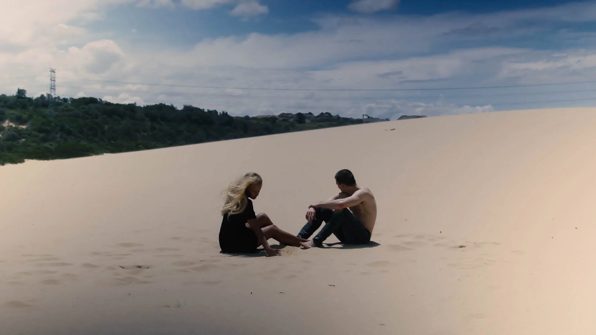 A man and woman sitting on a sandy beach, facing each other with their knees bent, in a secluded beach scene with cloudy sky and distant green hills.
